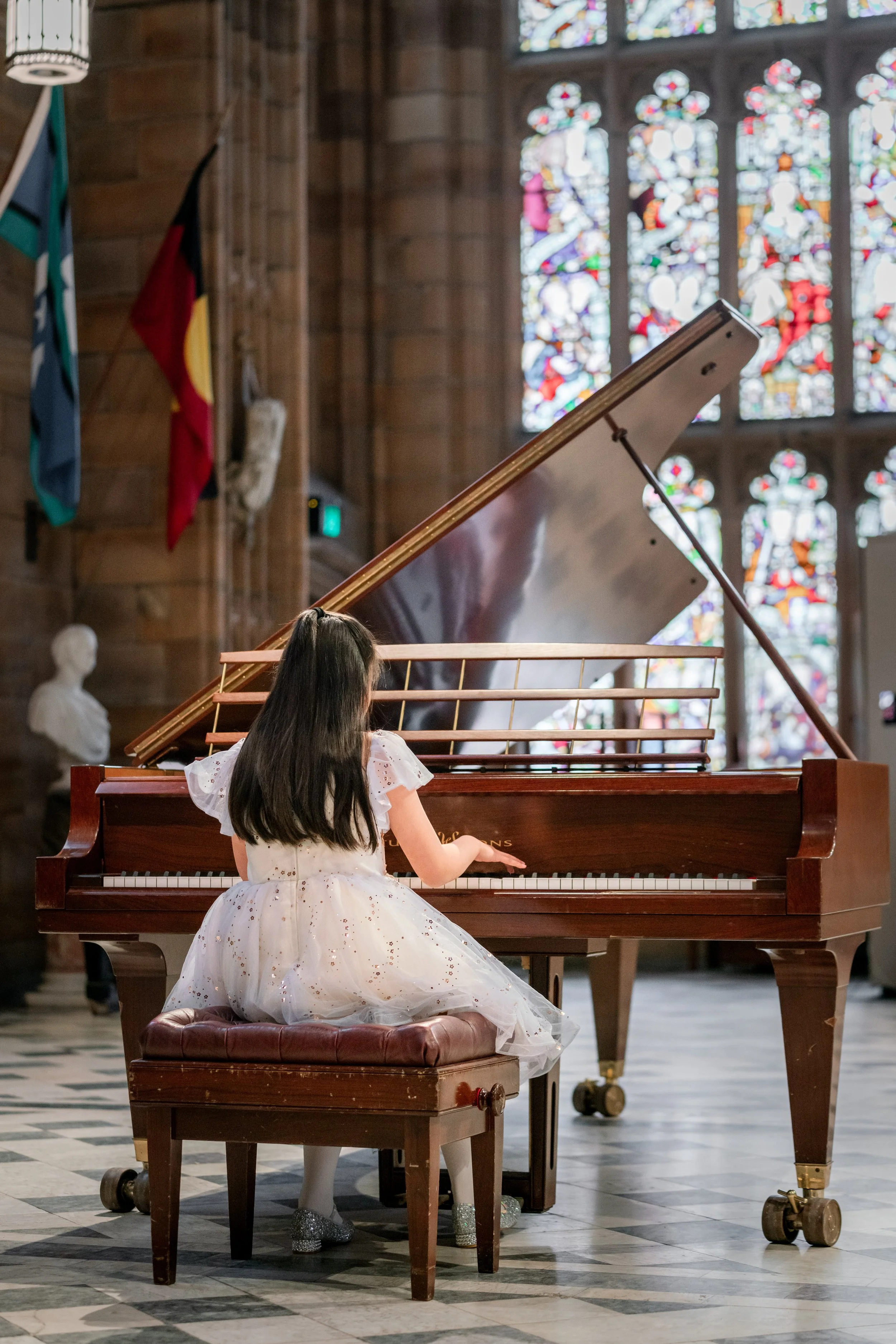A young girl in a white dress sitting at a grand piano in a church with stained glass windows.