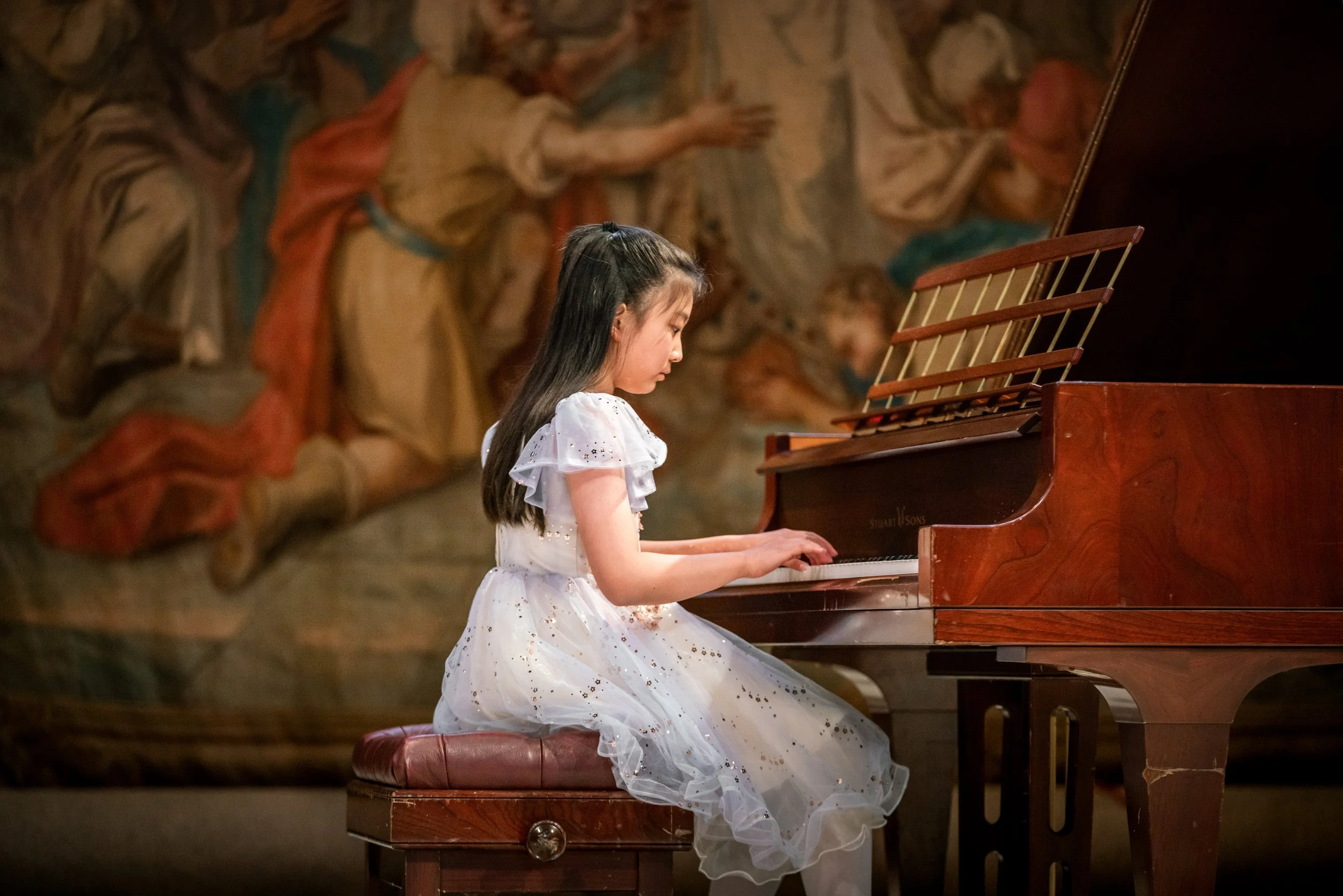 A young girl in a white dress playing a grand piano with a painted mural in the background.