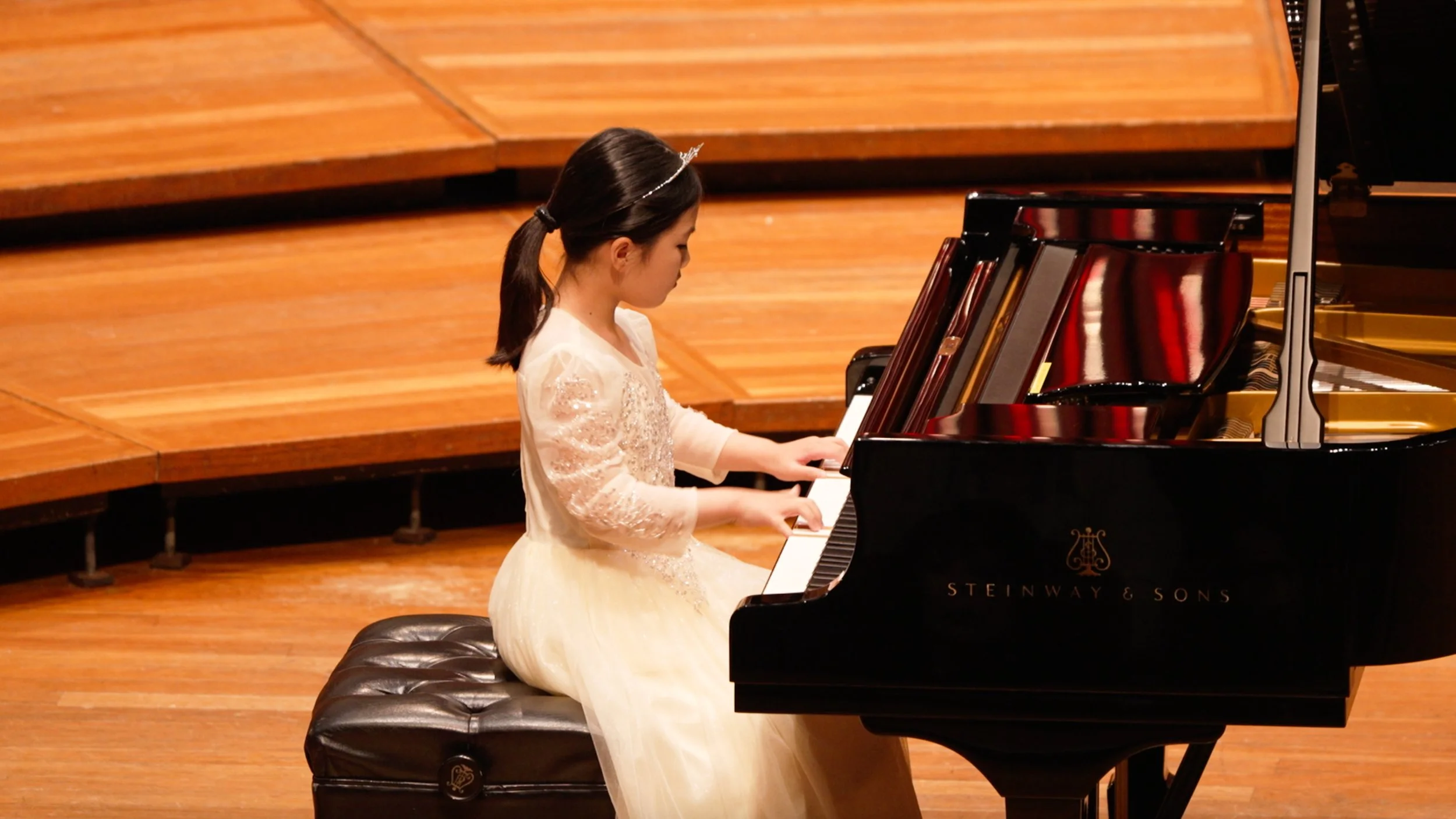 Young girl in a white dress playing a grand piano on a stage with wooden flooring