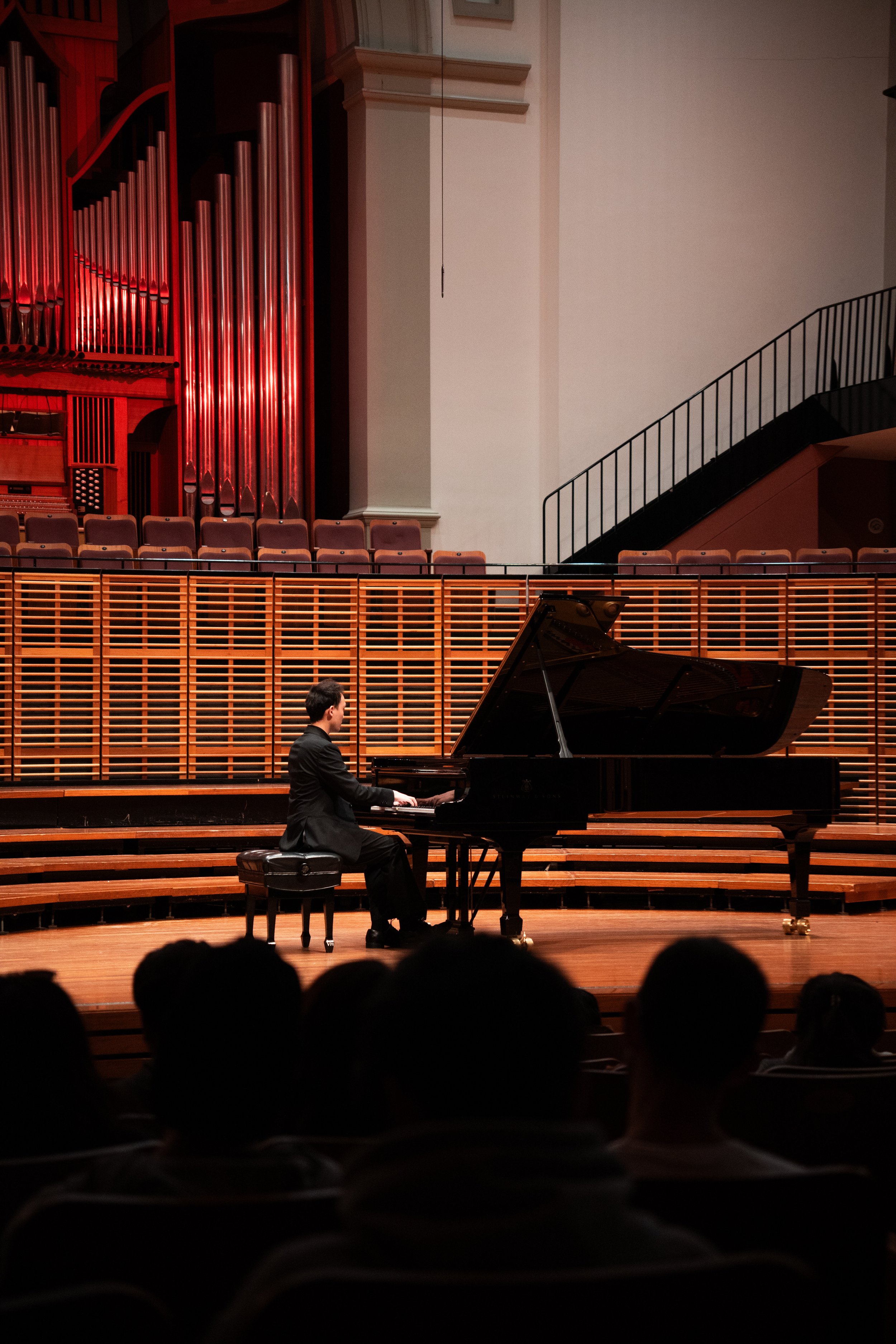 A man playing a grand piano on a concert hall stage, with audience members seated in front of him and a large pipe organ in the background.