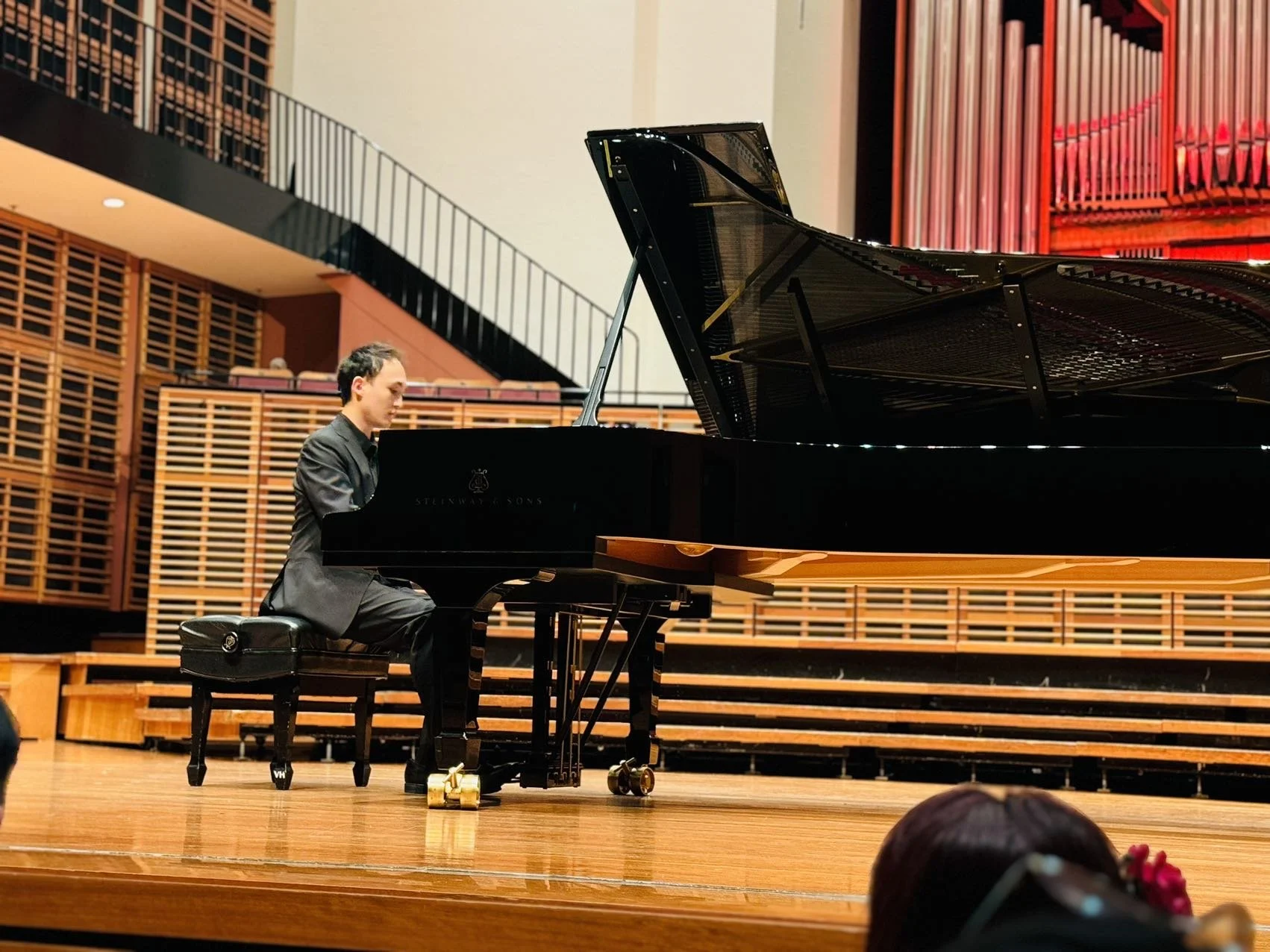 A man in formal attire playing a grand piano on a stage in a concert hall.
