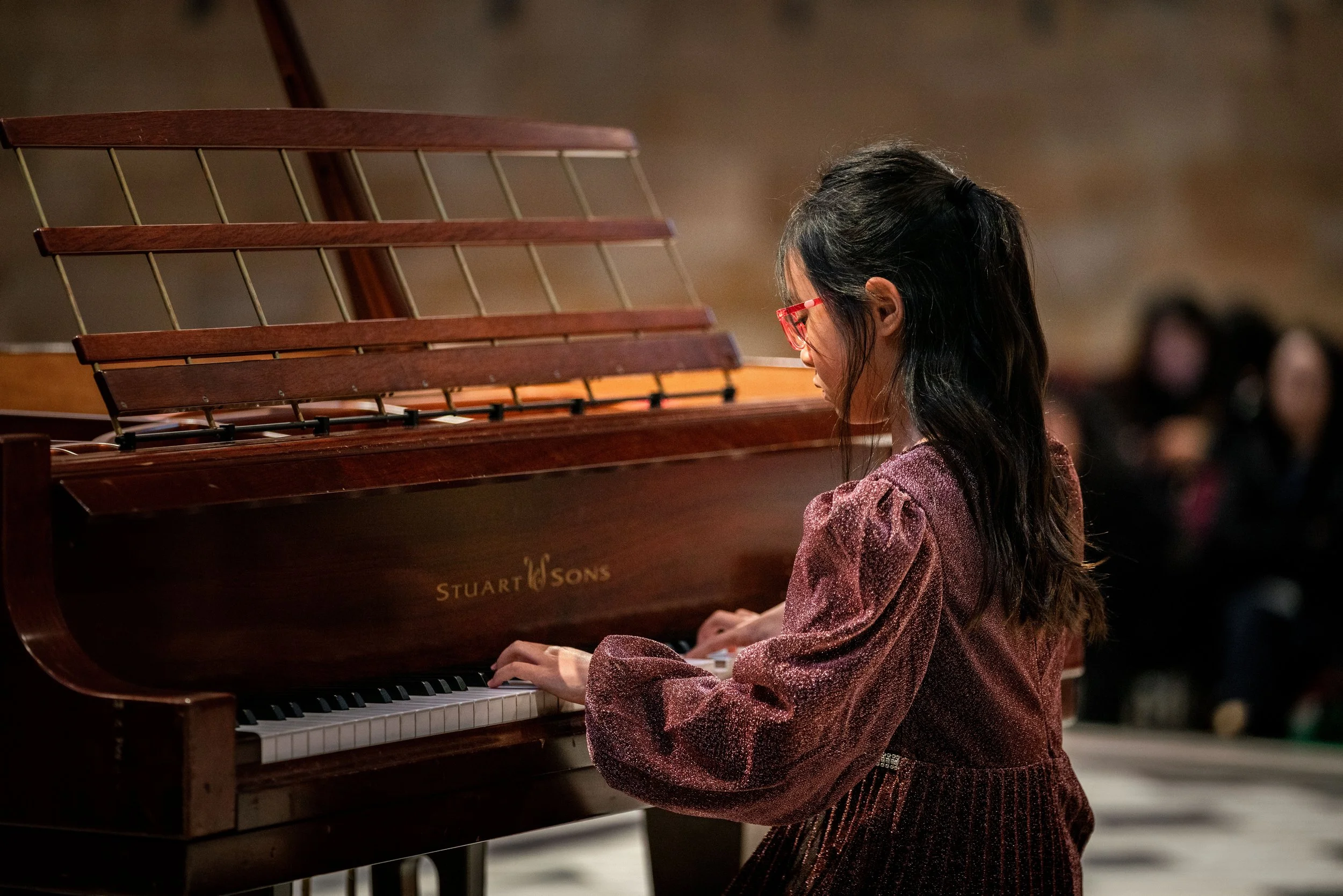 A young girl with long dark hair and red glasses playing a wooden upright piano in a dimly lit room with a blurred audience in the background.