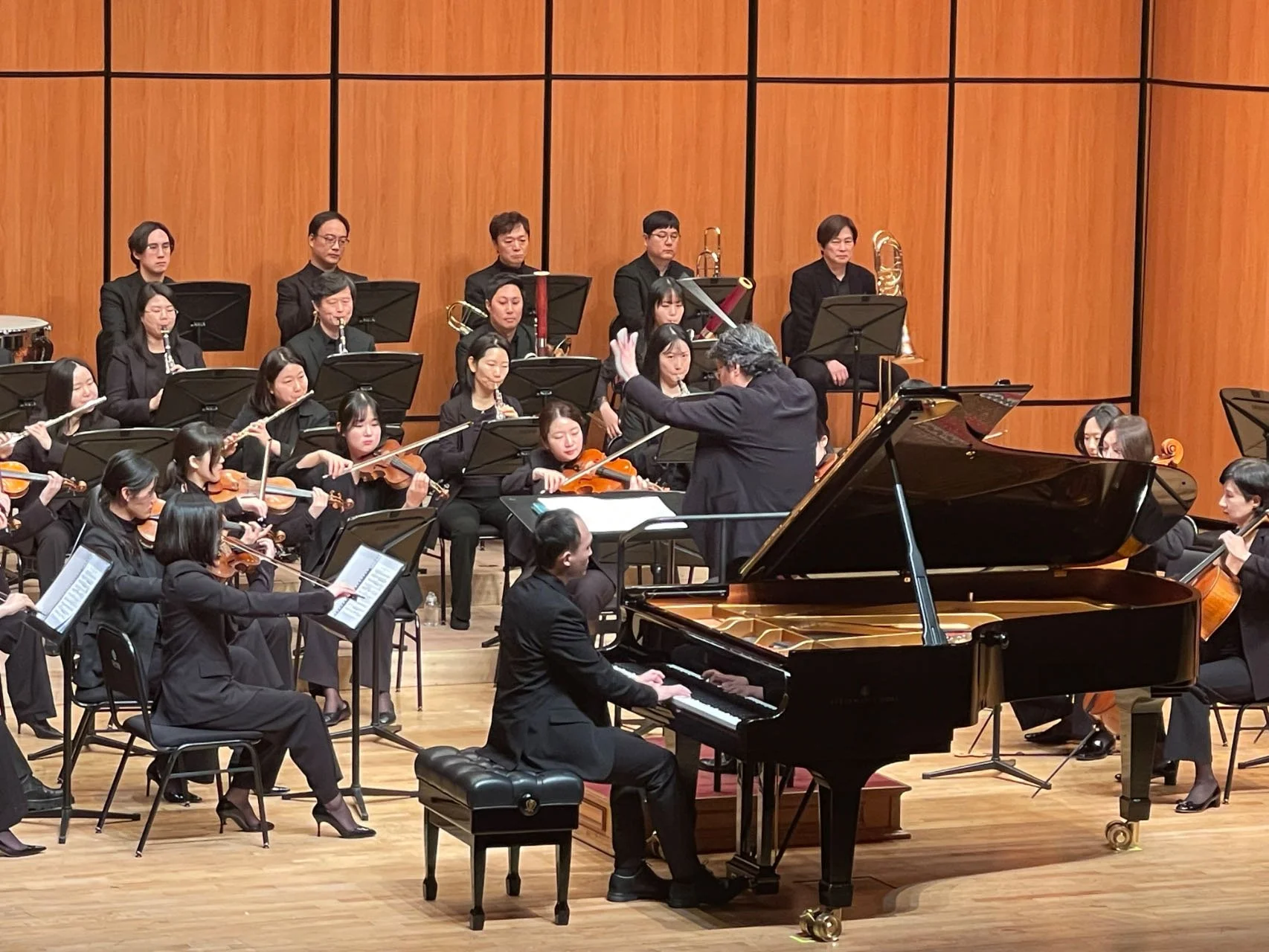 An orchestra performing on stage with musicians playing various instruments, including violins, and a pianist at a grand piano, led by a conductor.