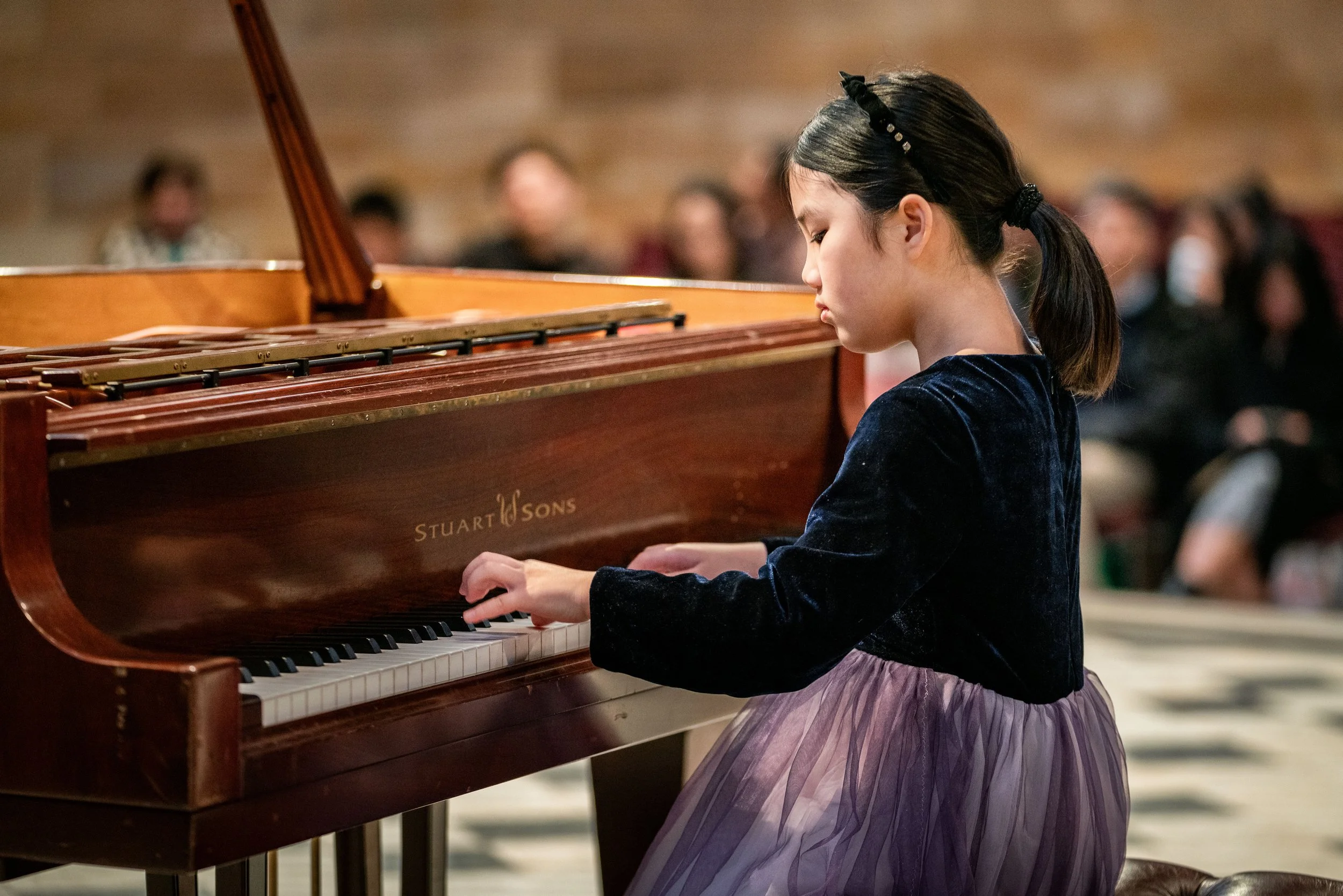 A young girl playing a wooden upright piano during a performance or concert with an audience in the background.