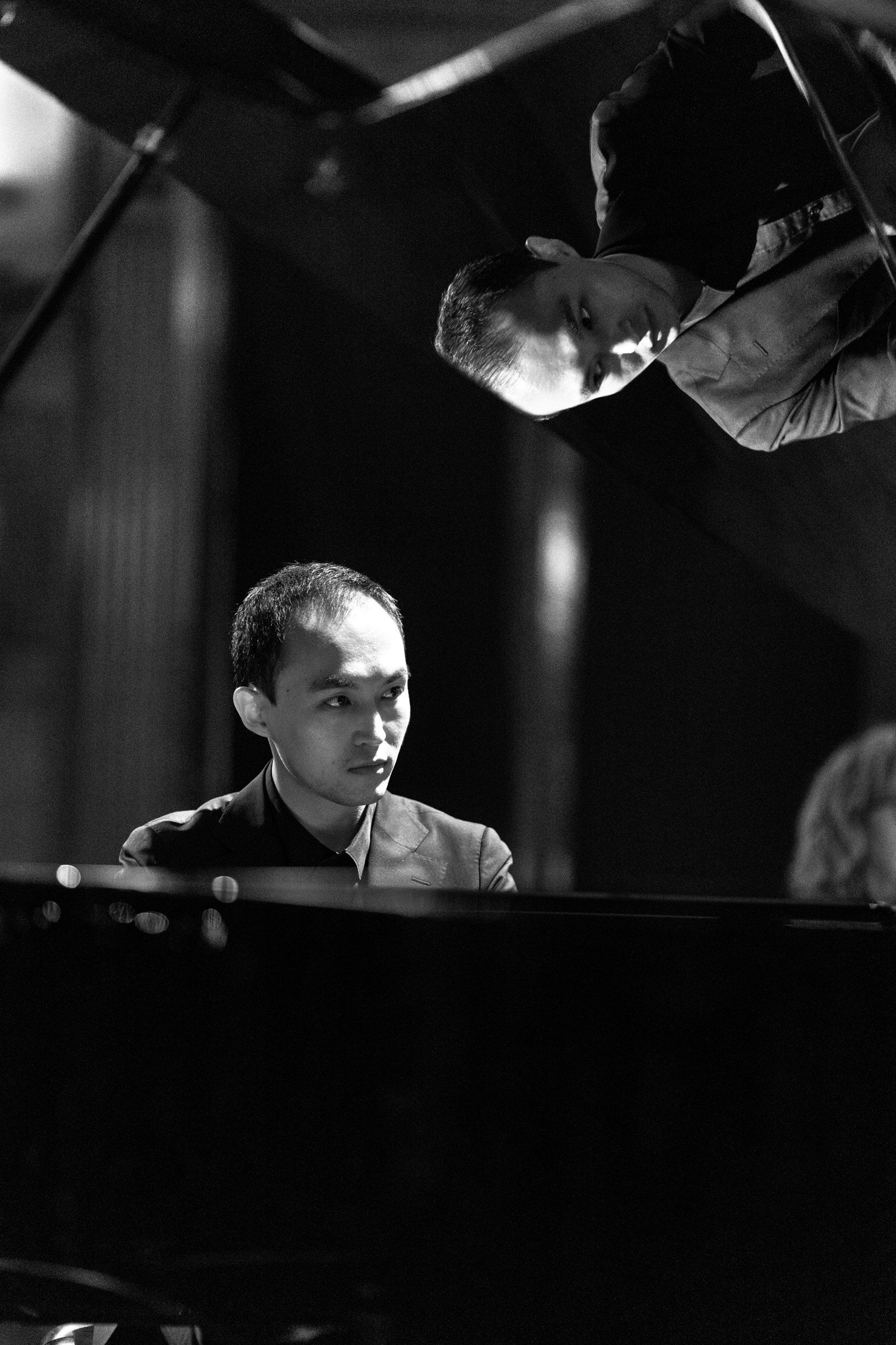 Two young men in formal attire playing a grand piano together in a dimly lit room.