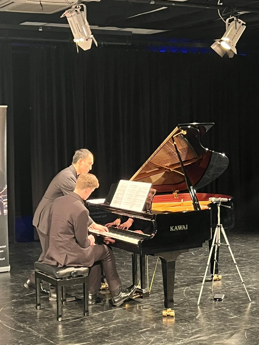 Two men in suits playing a grand piano on a stage with black curtains, with sheet music open on the piano.