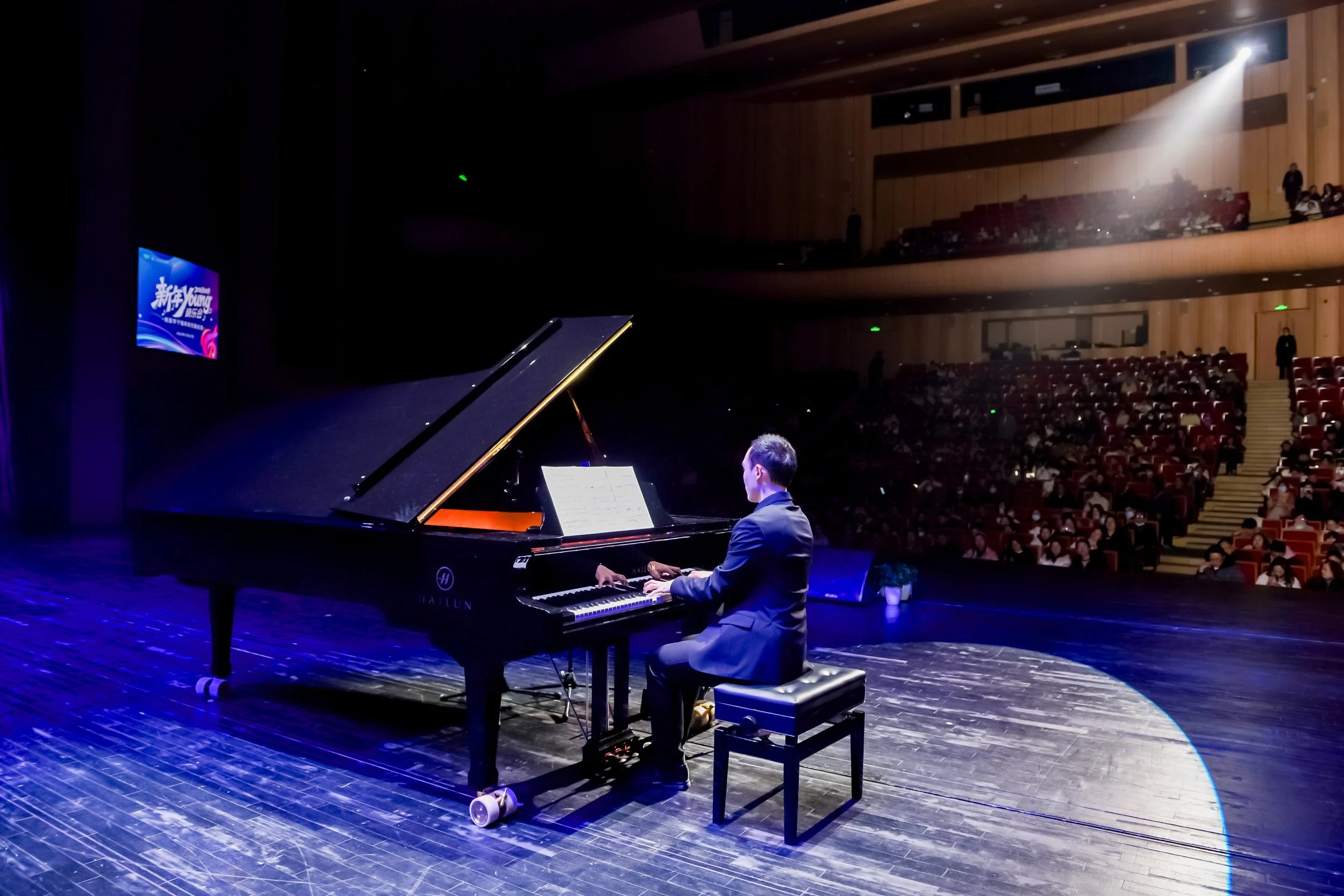 A man in a suit playing the piano on a stage in a concert hall with an audience watching.