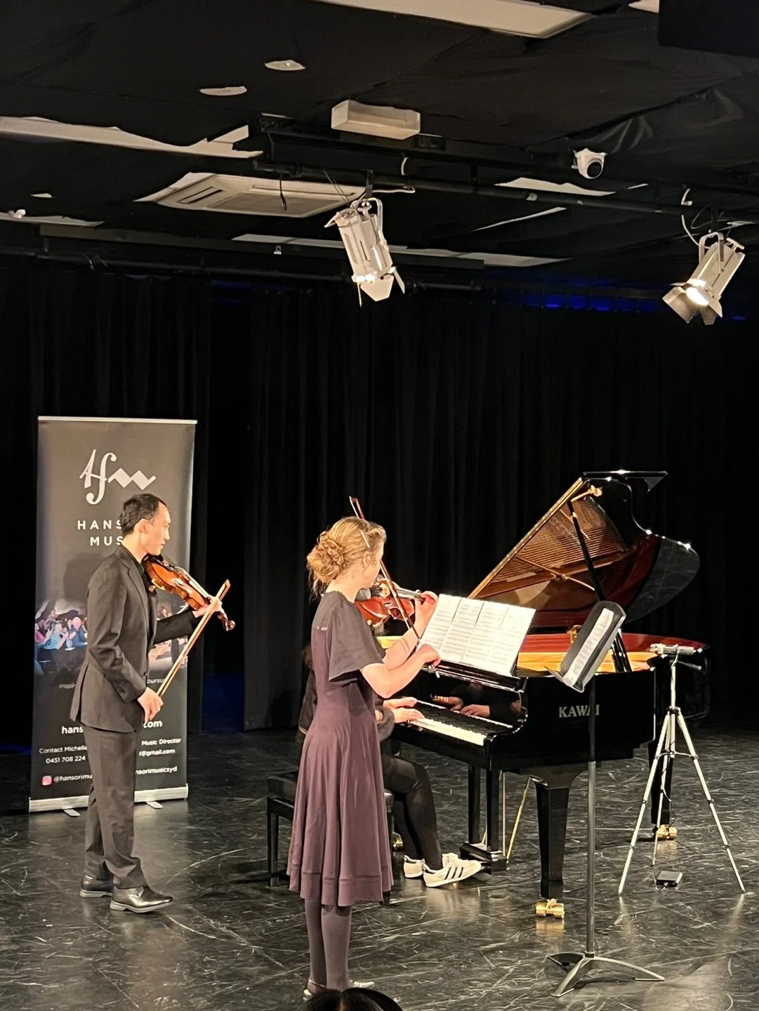 Three musicians performing on stage: a violinist standing to the left, a pianist seated at a grand piano, and a violinist standing to the right, all focused on their music.