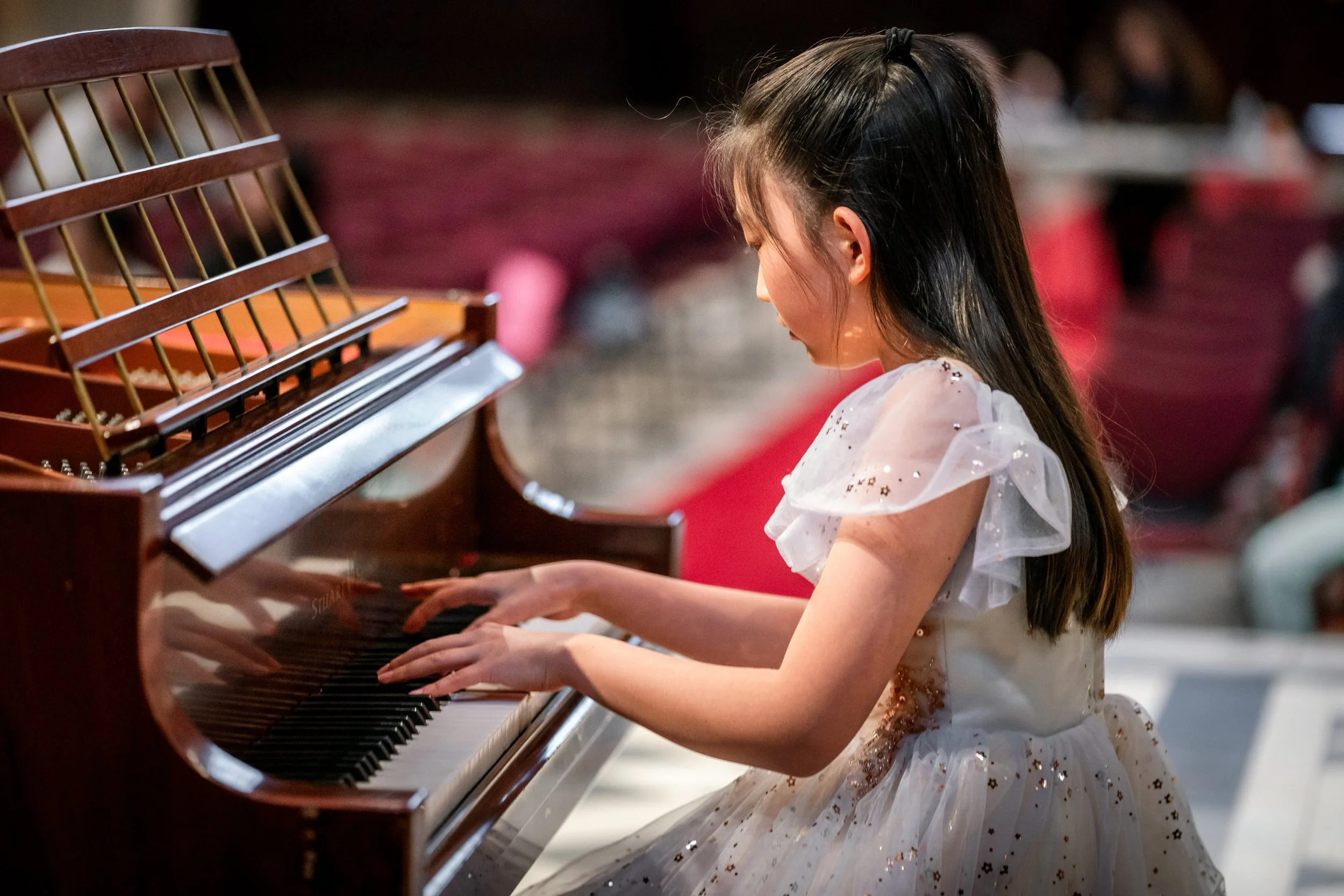 Young girl playing the piano, dressed in a white dress with ruffled sleeves and gold accents, sitting at a grand piano in a performance setting.