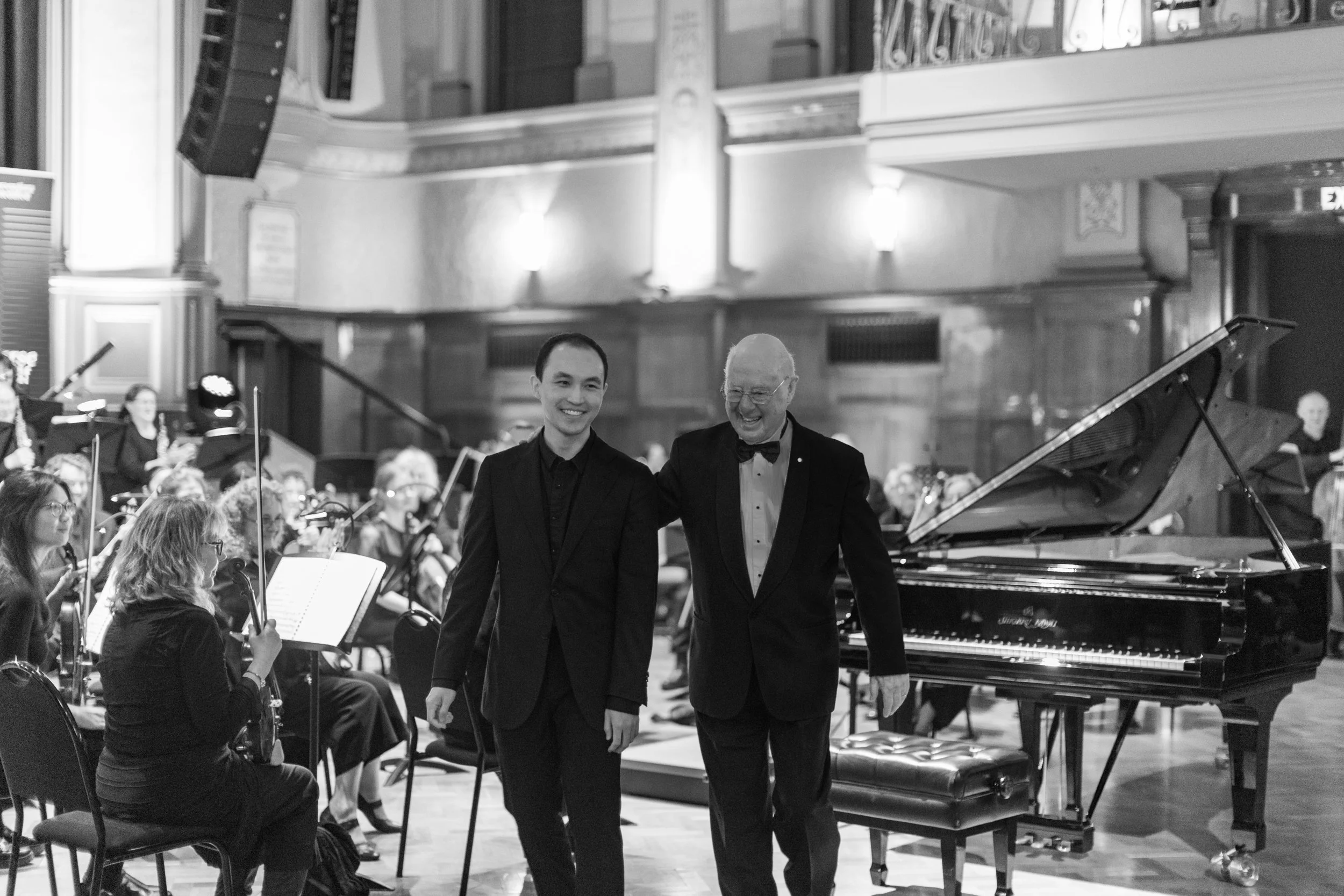 Two men in formal tuxedos standing and smiling in front of an orchestra during a concert in a grand hall, with a grand piano beside them.