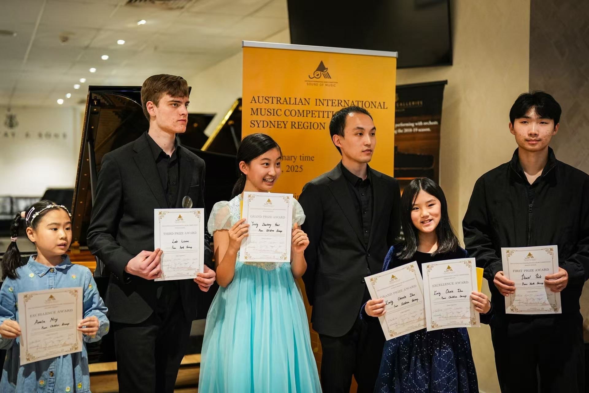 Six young people standing on a stage holding certificates at an award ceremony for a music competition in Sydney. Background features a piano and a yellow banner reading 'Australian International Music Competition, Sydney Region'.
