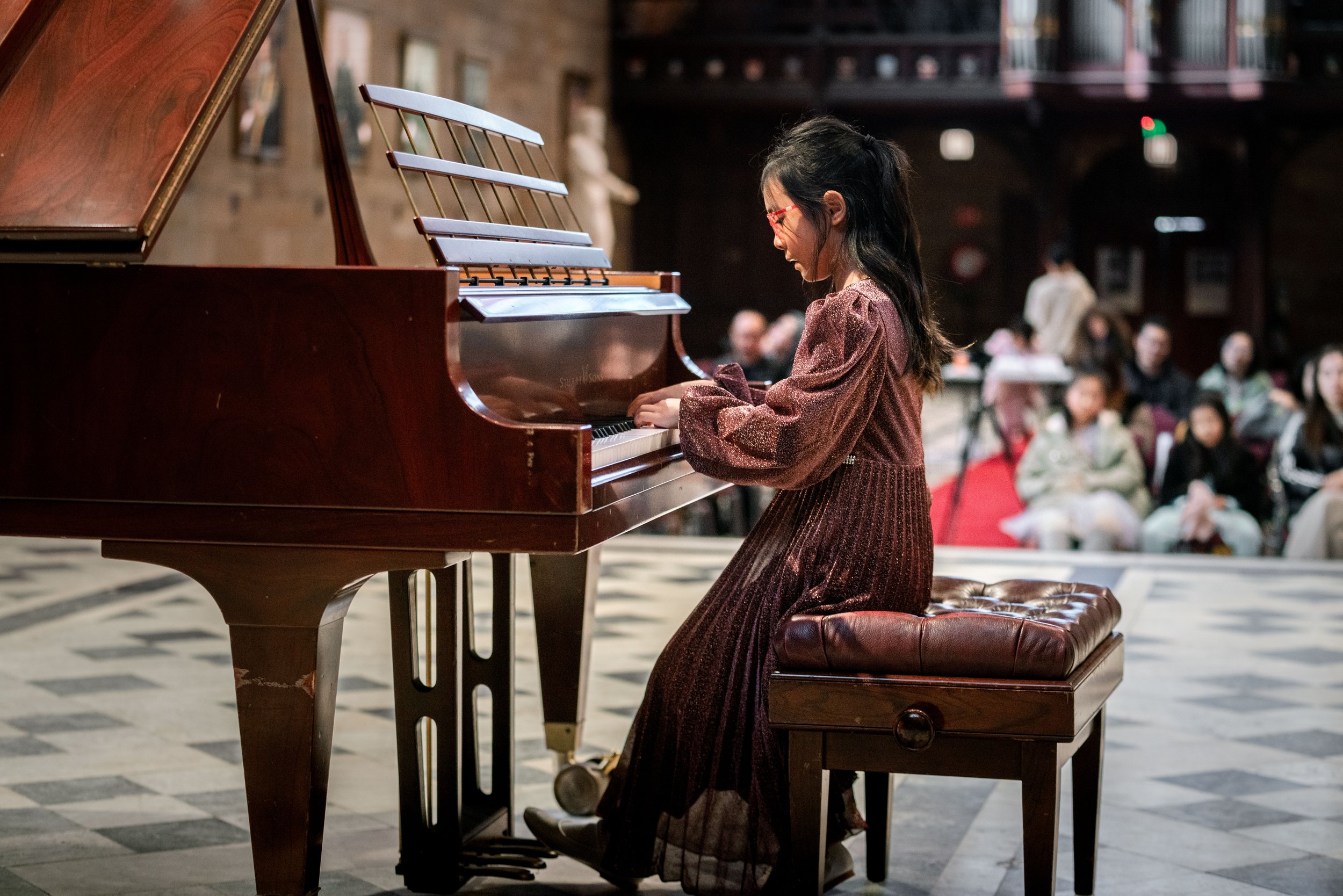 A young woman wearing glasses and a long dress playing a grand piano in a warmly lit indoor concert hall with an audience seated in the background.