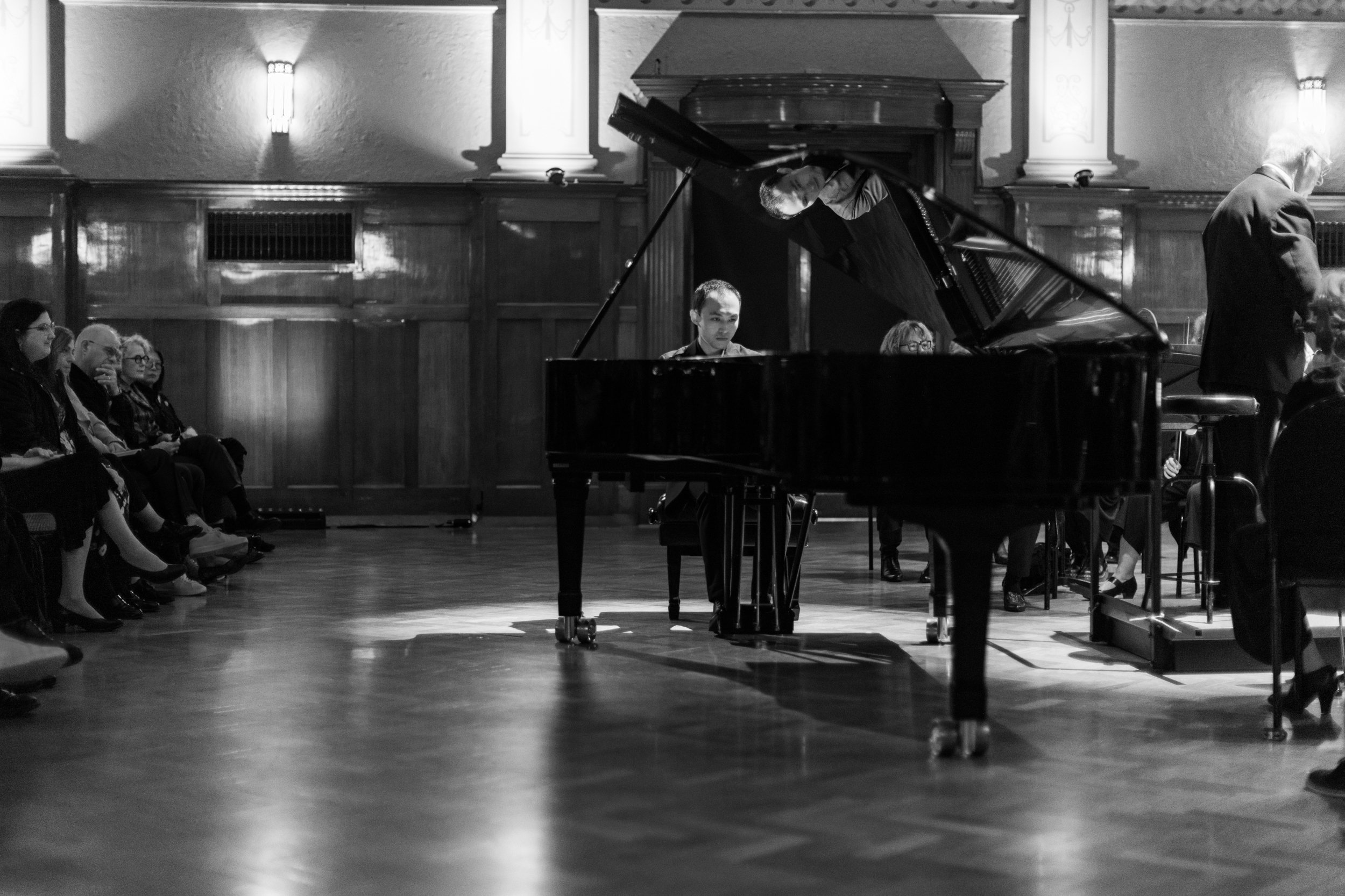Black and white photo of a music recital in a concert hall, with a pianist playing on a grand piano surrounded by audience members seated on chairs.