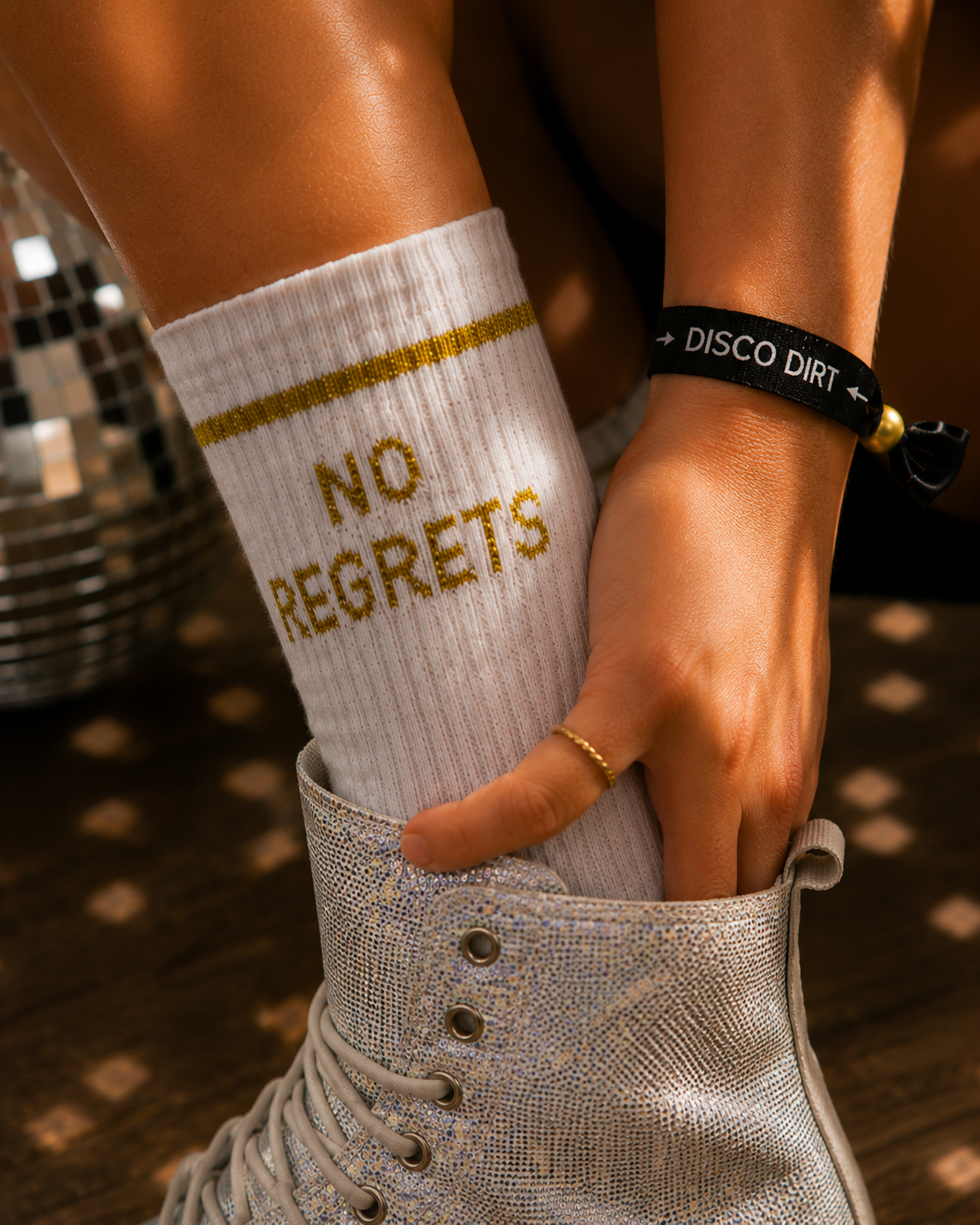 Close-up of a person's foot wearing a sparkly silver boot and a white sock with yellow lettering that says 'NO REGRETS'. The person is also wearing a black bracelet that says 'DISCO DIRT' and a gold ring.