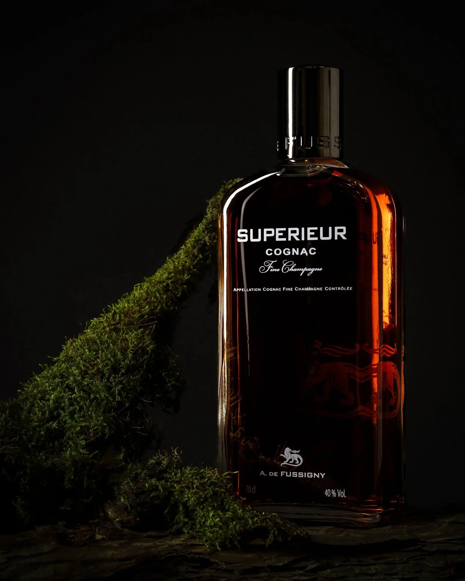Eine Flasche Cognac mit schwarzem Etikett, daneben Moos auf einer Holzoberfläche gegen schwarzen Hintergrund.