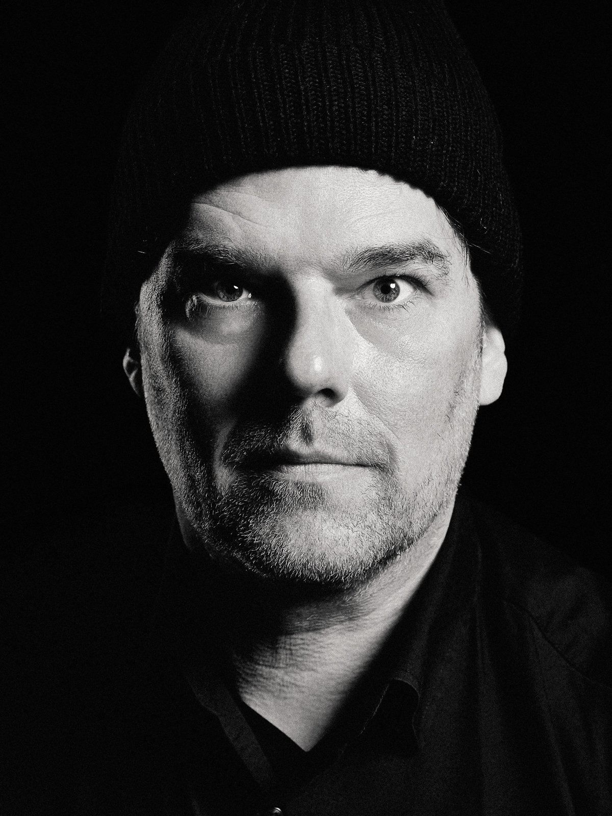 Schwarz-weiß Porträt eines Mannes mit grauem Bart, schwarzer Kappe und dunklem Hemd, Blick direkt in die Kamera.