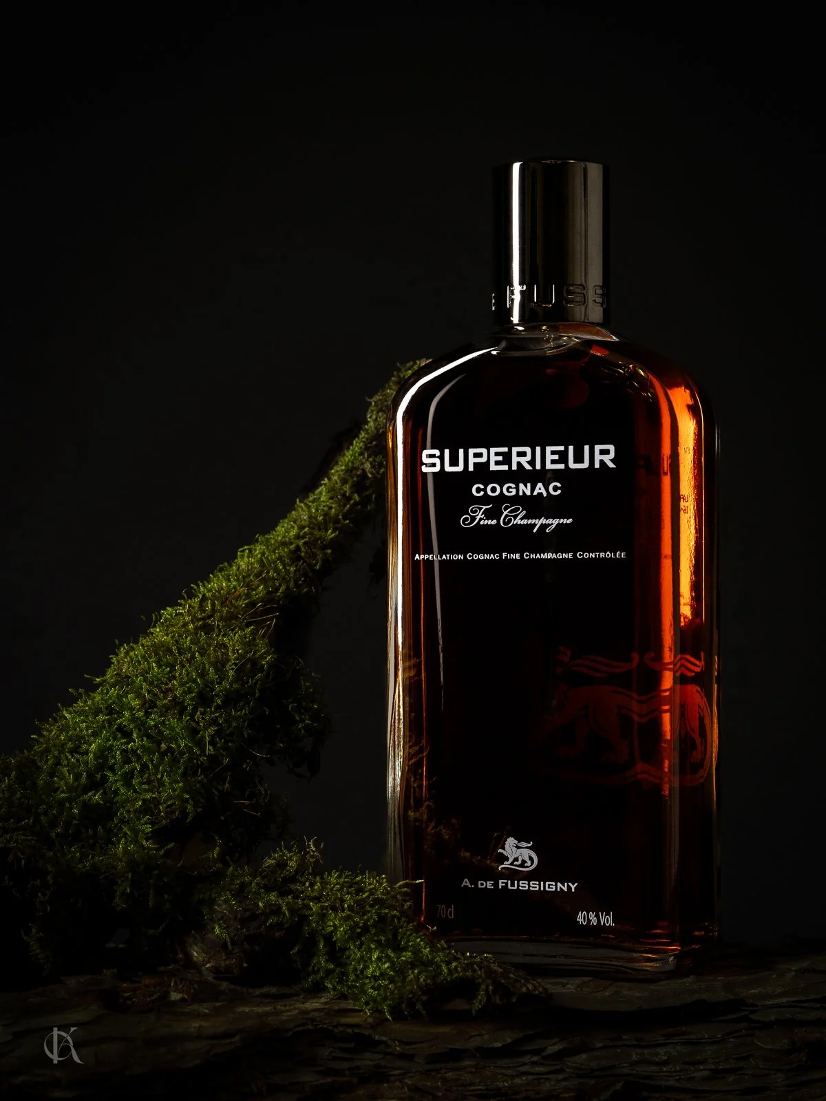 Eine dunkle Flasche Cognac mit weißem Aufdruck steht auf einem Holzstück neben einem grünen Moosstängel auf schwarzem Hintergrund.