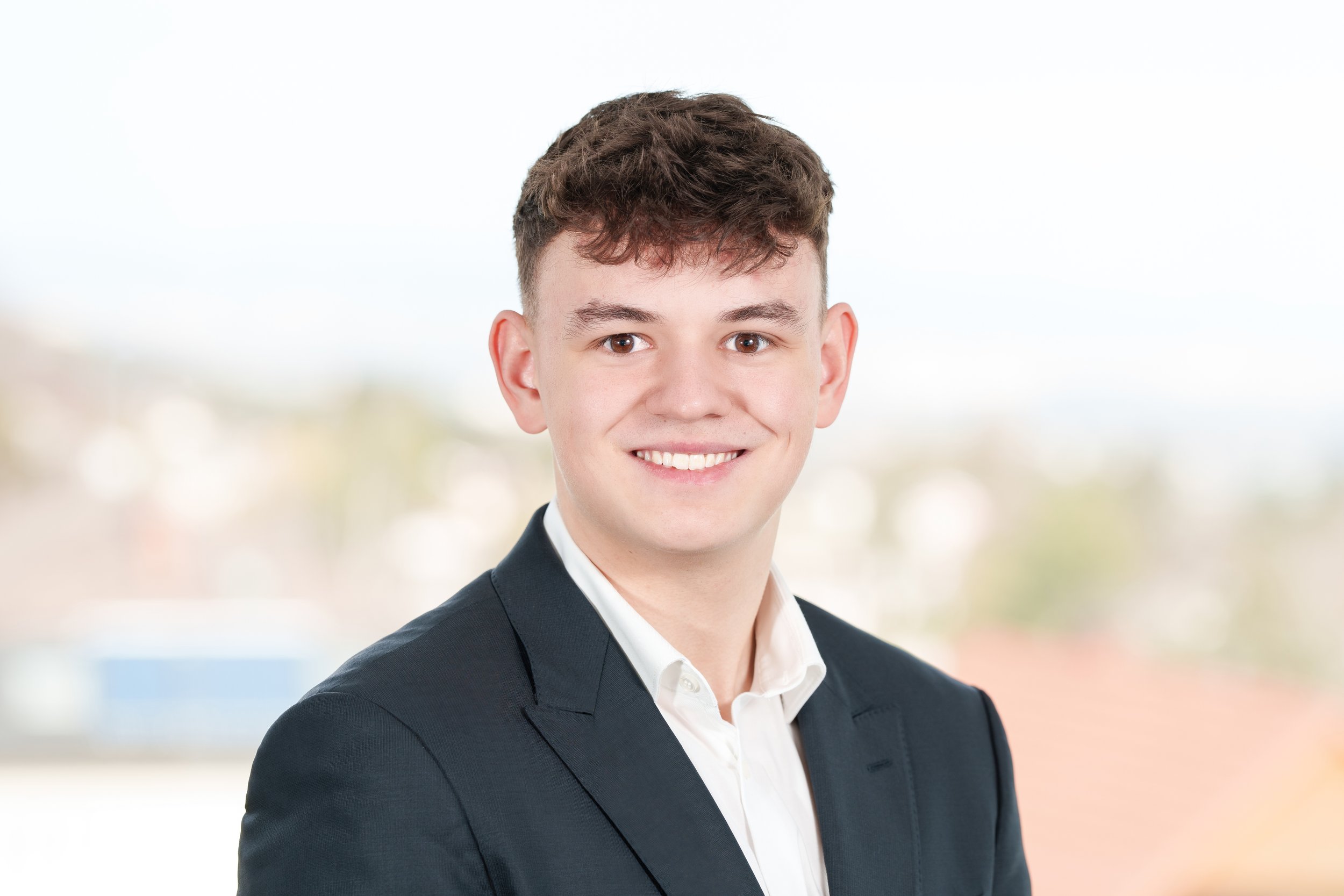 Junger Mann in formellem Anzug mit weißem Hemd, lächelnd vor hellem Hintergrund.