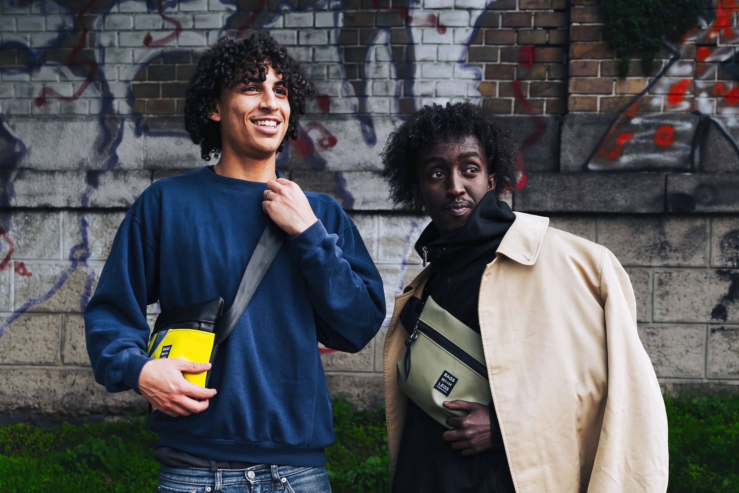 Zwei junge Männer vor einer Wand mit Graffiti, lächelnd und die Taschen umhängen.
