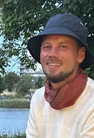 Mand med stråhat og tørklæde, stående ved vand med grønne træer og bybygninger i baggrunden.