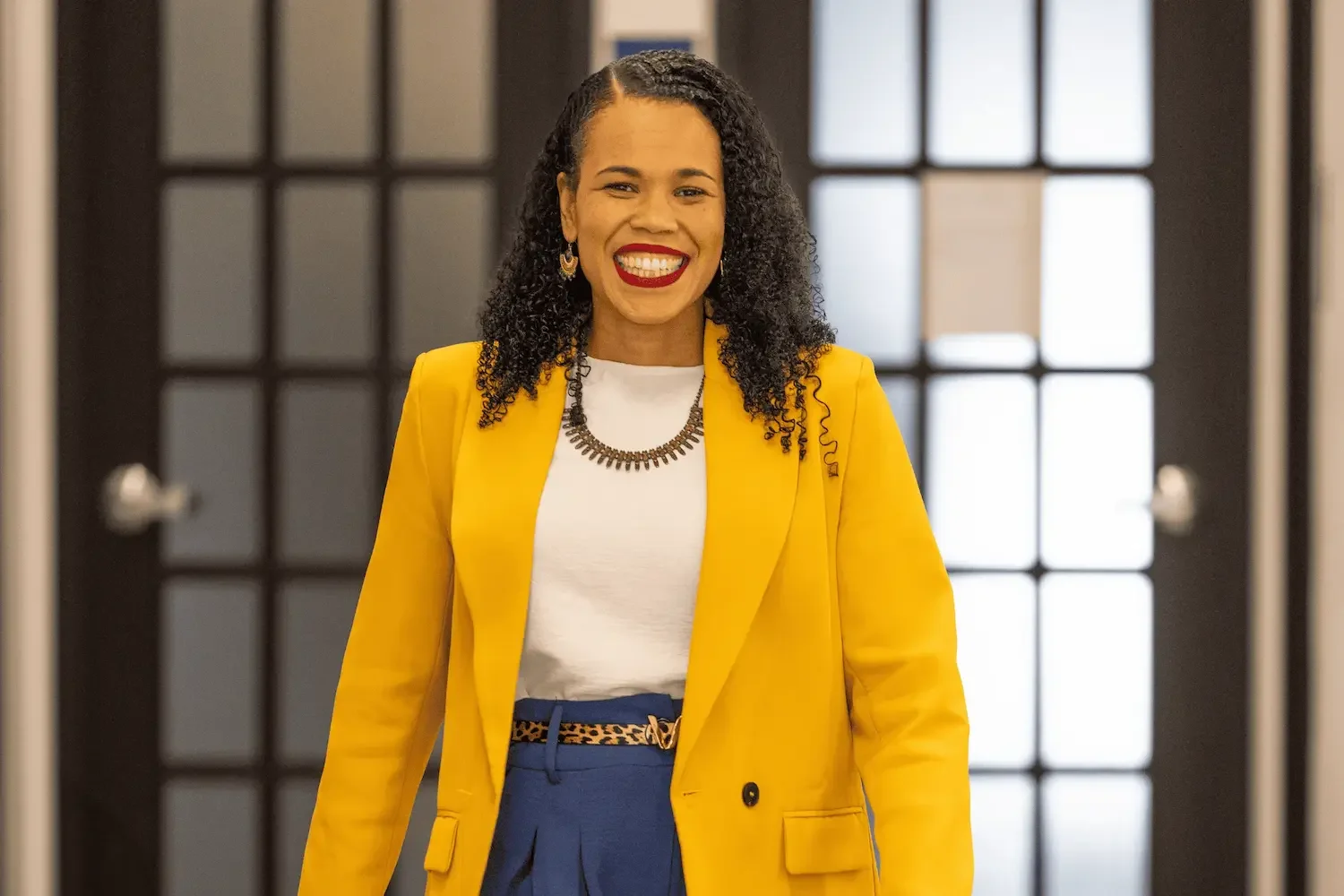 Sandrine Edmond Une femme souriante vêtue d'un blazer jaune, d'un t-shirt blanc, d'une ceinture à motif léopard et d'un collier noir, dans un intérieur moderne.