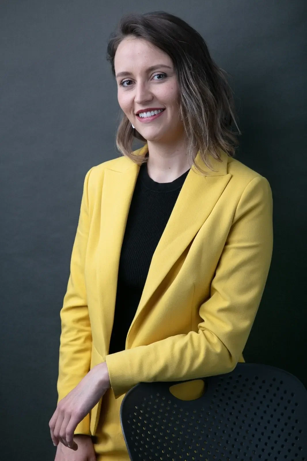 Femme souriante portant un blazer jaune et un t-shirt noir, debout contre un fond gris foncé, avec une chaise noire en premier plan.