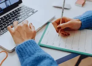 Personne écrivant dans un carnet à côté d'un ordinateur portable sur un bureau.