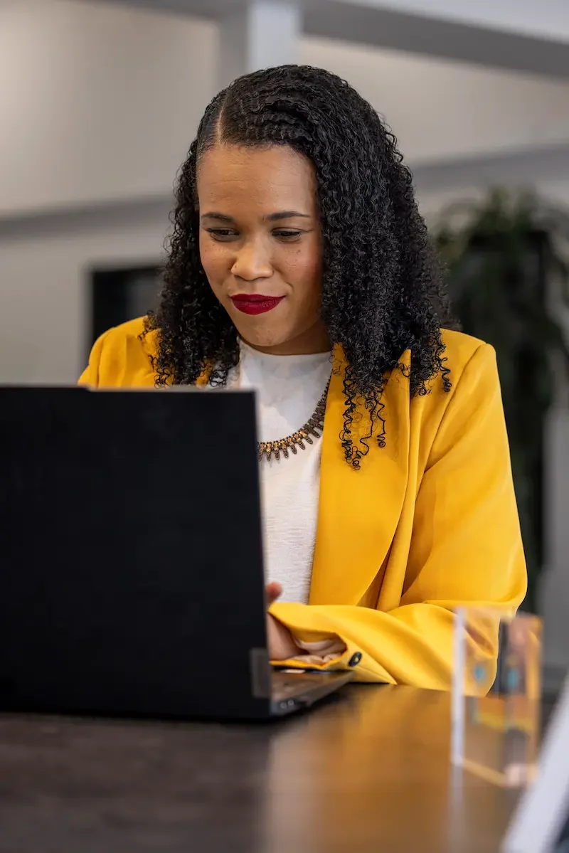 Sandrine Edmond  : Femme assise devant un ordinateur portable, portant une veste jaune, dans un environnement professionnel.