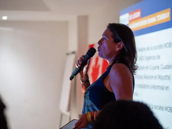 Une femme parle dans un micro lors d'une présentation ou conférence, devant un écran affichant du texte en français.