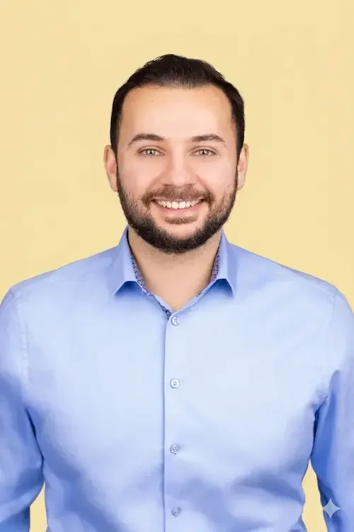 photo d'un consultant de Soleil Immigration :  Un homme souriant avec une barbe, portant une chemise bleu clair, photo en studio contre un fond jaune pastel.