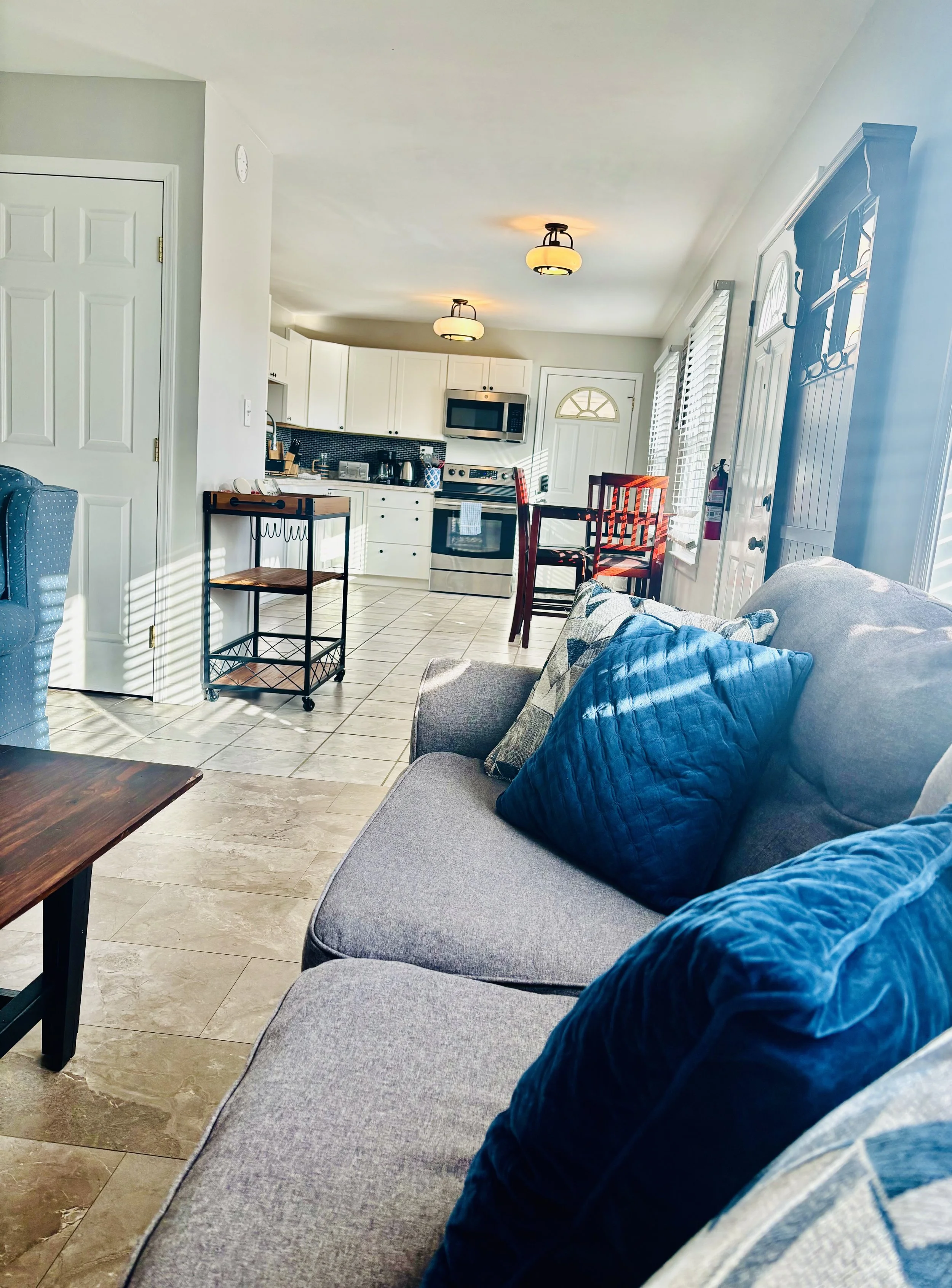 Living room area with a gray couch and blue pillows in the foreground, kitchen with white cabinets, a microwave, and a tiled backsplash in the background, sunlight streaming in through large windows.