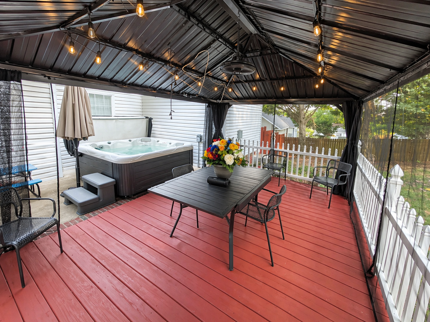 Covered patio with red wooden flooring, string lights, a hot tub, a black dining table with a flower arrangement, and several black chairs.