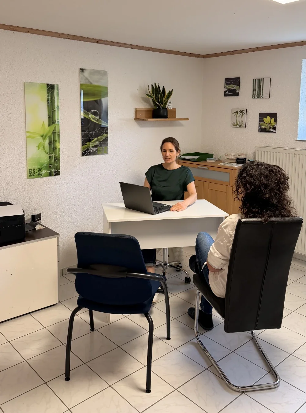 Heilpraktikerin Beatrice Bayer sitzt lächelnd an einem weißen Schreibtisch mit Laptop und berät eine Patientin in ihrer hellen Praxis. Die ruhige Atmosphäre wird durch grüne Naturmotive an den Wänden und eine Zimmerpflanze unterstrichen.