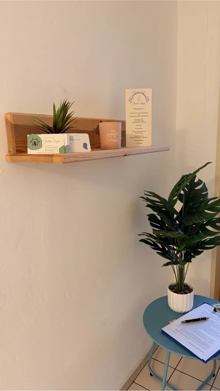 Ein helles Holzregal an einer cremefarbenen Wand präsentiert Visitenkarten und eine Infotafel der Naturheilkundepraxis von Heilpraktikerin Beatrice Bayer. Eine kleine Sukkulente und eine lachsfarbene Kerze mit der Aufschrift „Take a moment to smile“