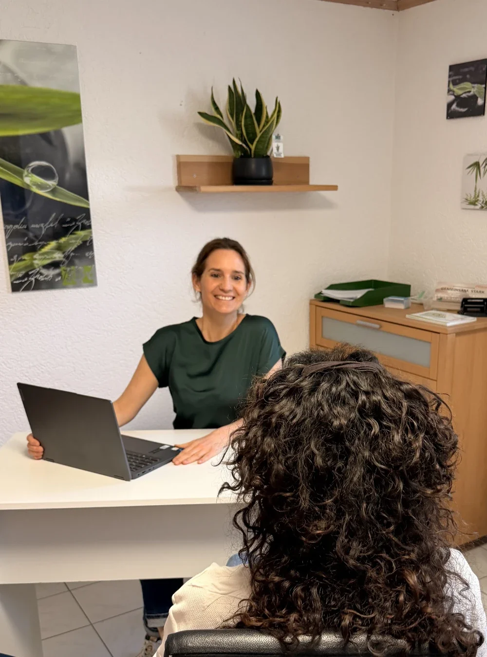 Heilpraktikerin Beatrice Bayer sitzt an ihrem Schreibtisch und lächelt ihre Patientin bei der Beratung strahlend an. Das helle Büro wirkt sehr persönlich und schafft eine vertrauensvolle Atmosphäre für das therapeutische Gespräch.