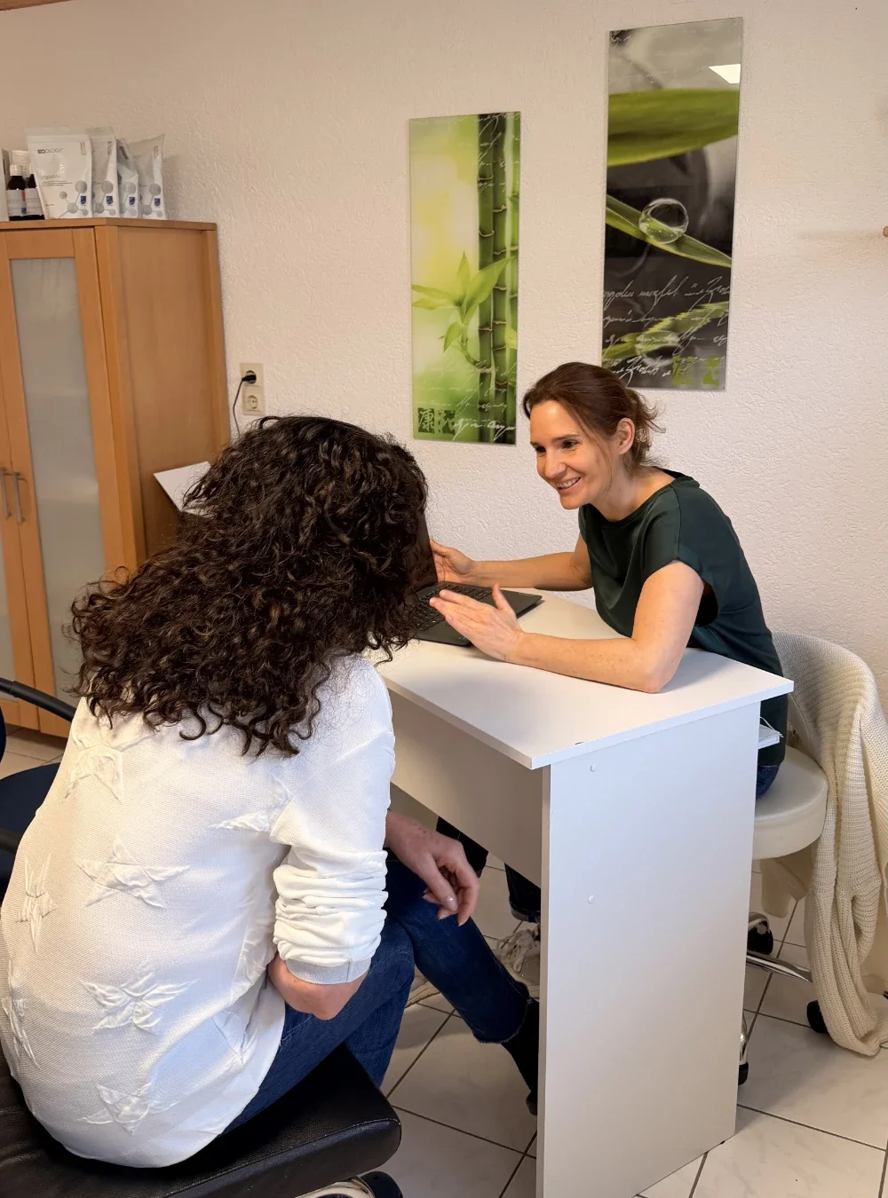 Heilpraktikerin Beatrice Bayer im freundlichen Beratungsgespräch mit einer Patientin in ihrer hellen Praxis. Sie sitzt lächelnd am weißen Schreibtisch vor einem Laptop, während im Hintergrund beruhigende Naturmotive die Atmosphäre unterstreichen.