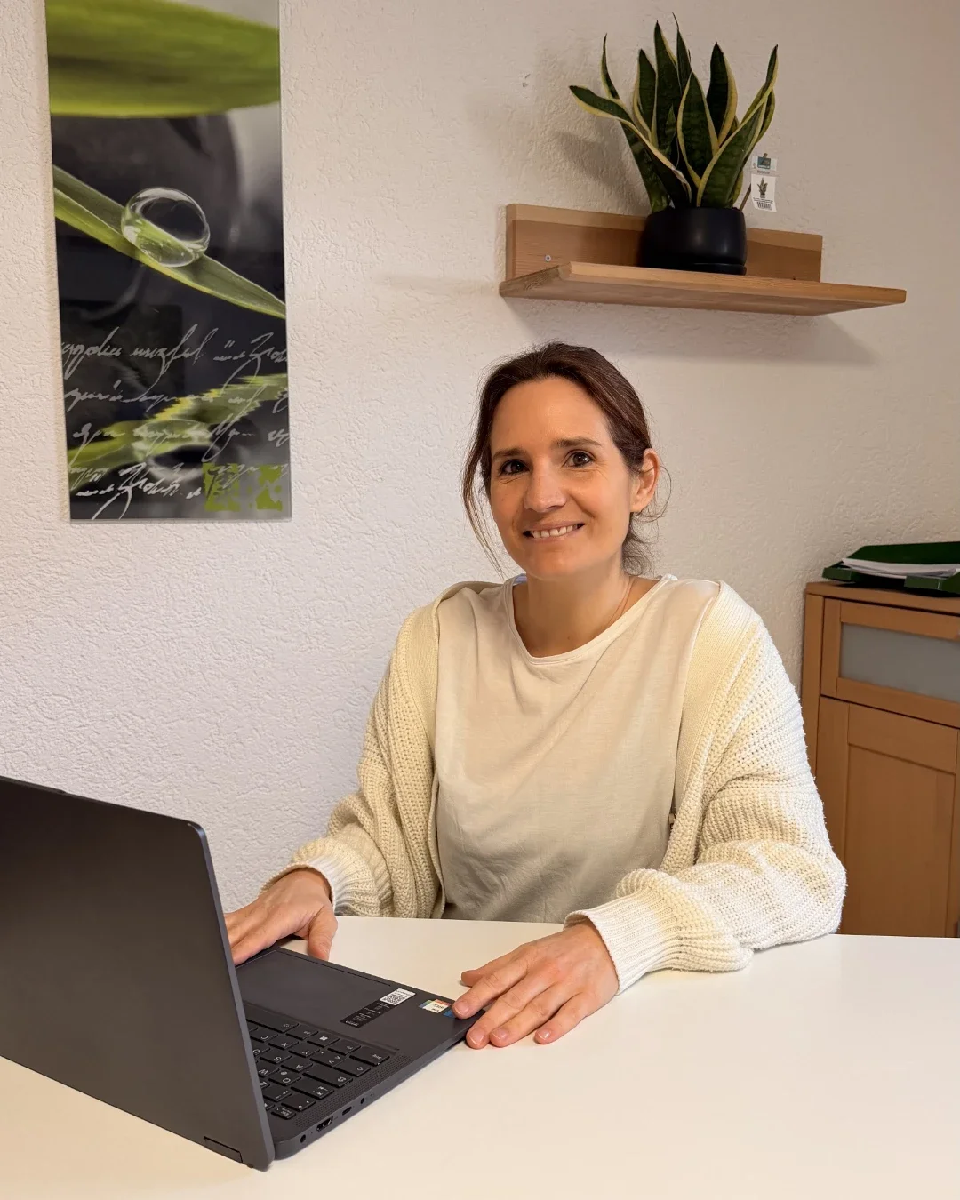 Heilpraktikerin Beatrice Bayer sitzt lächelnd an einem weißen Schreibtisch vor ihrem geöffneten Laptop und blickt freundlich direkt in die Kamera. Die helle, einladende Büroumgebung wird durch eine Grünpflanze auf einem Holzregal und ein harmonisches