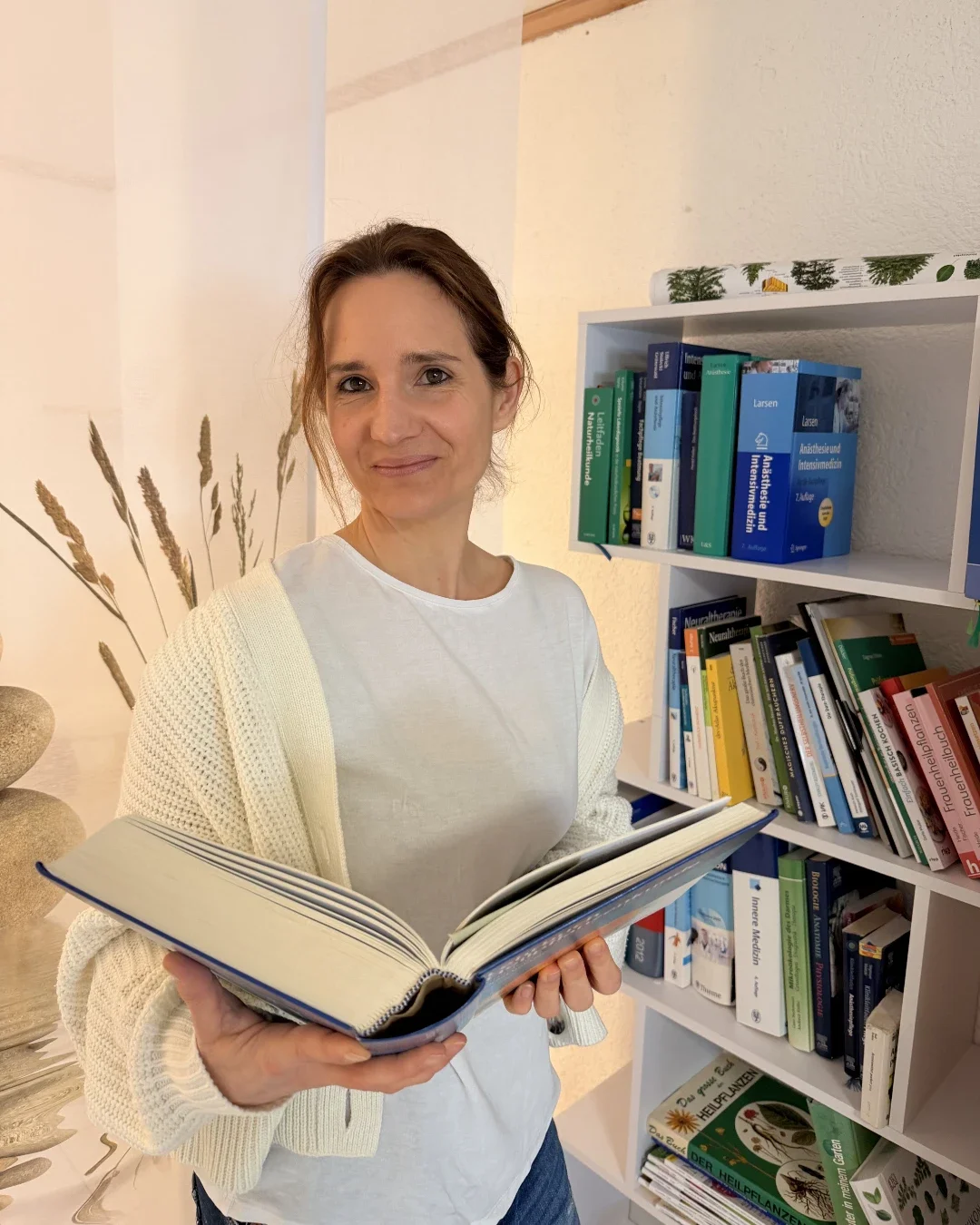 Heilpraktikerin Beatrice Bayer steht lächelnd in ihrer Praxis und hält ein aufgeschlagenes Fachbuch in den Händen. Im Hintergrund ist ein weißes Regal mit medizinischer Literatur und Büchern über Naturheilkunde zu sehen, was ihre fachliche Kompetenz