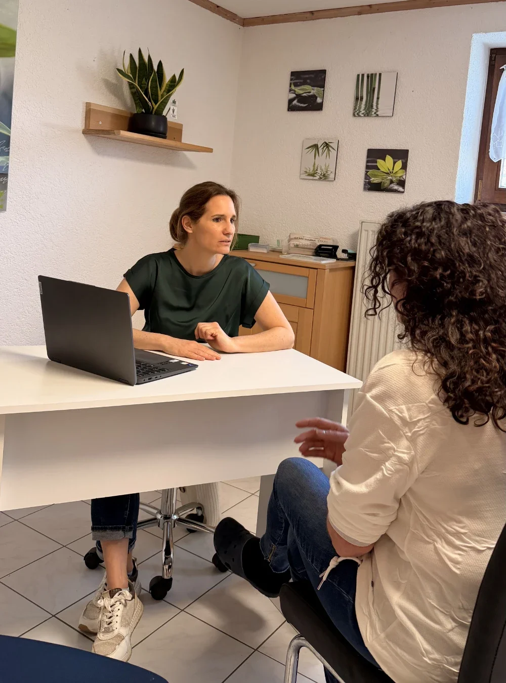 Heilpraktikerin Beatrice Bayer sitzt lächelnd an einem hellen Schreibtisch vor ihrem Laptop und berät eine Patientin mit dunklen Locken, die im Vordergrund Rücken an Rücken zum Betrachter sitzt. Die helle Praxis ist mit einer grünen Pflanze auf einem