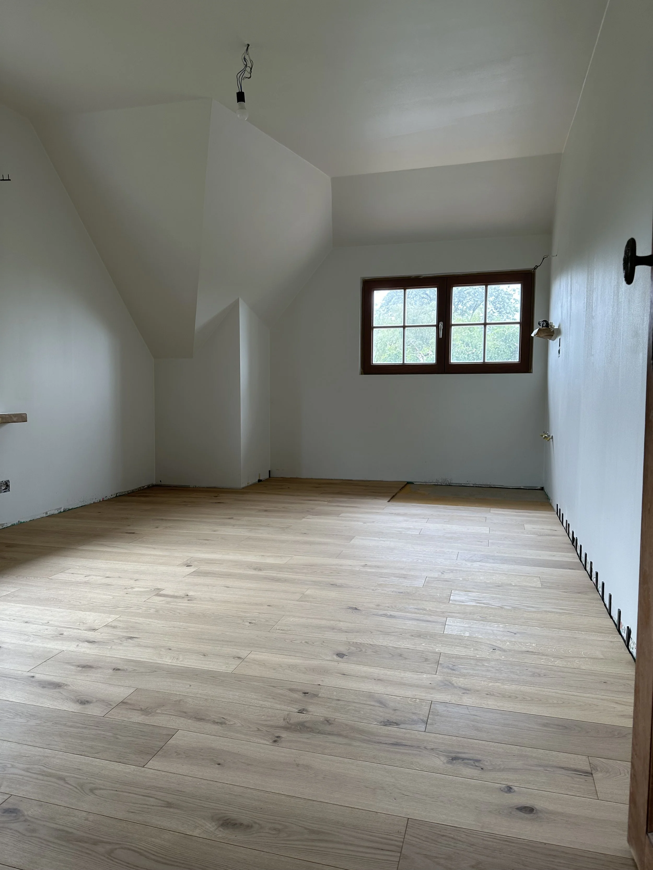 Empty room with white walls, light wood flooring, a window, and uninstalled light fixture hanging from the ceiling.
