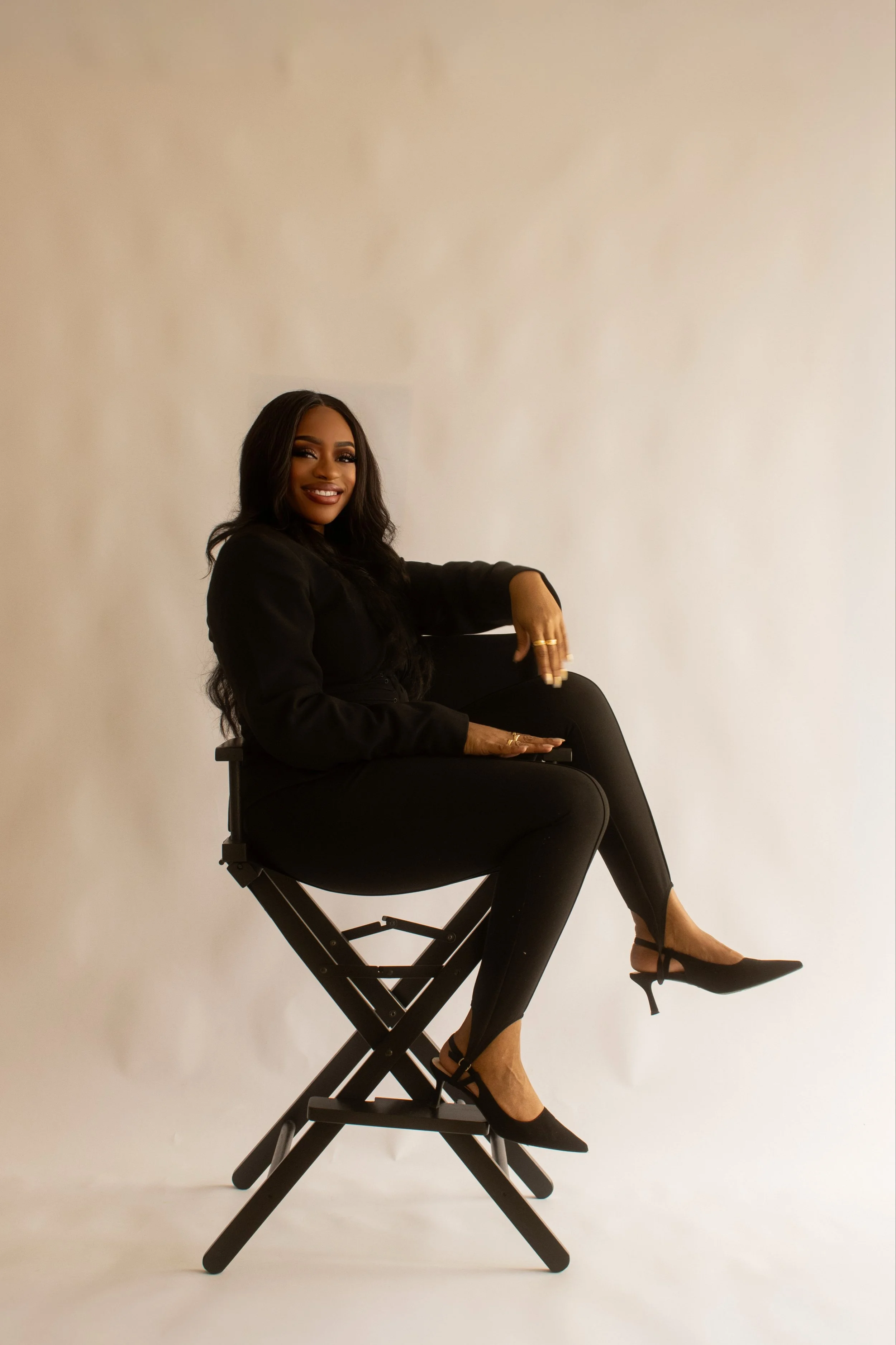 A woman with long dark hair and a bright smile, dressed in a black outfit, sitting on a director's chair against a plain white background. She is wearing black high heels and gold jewelry.