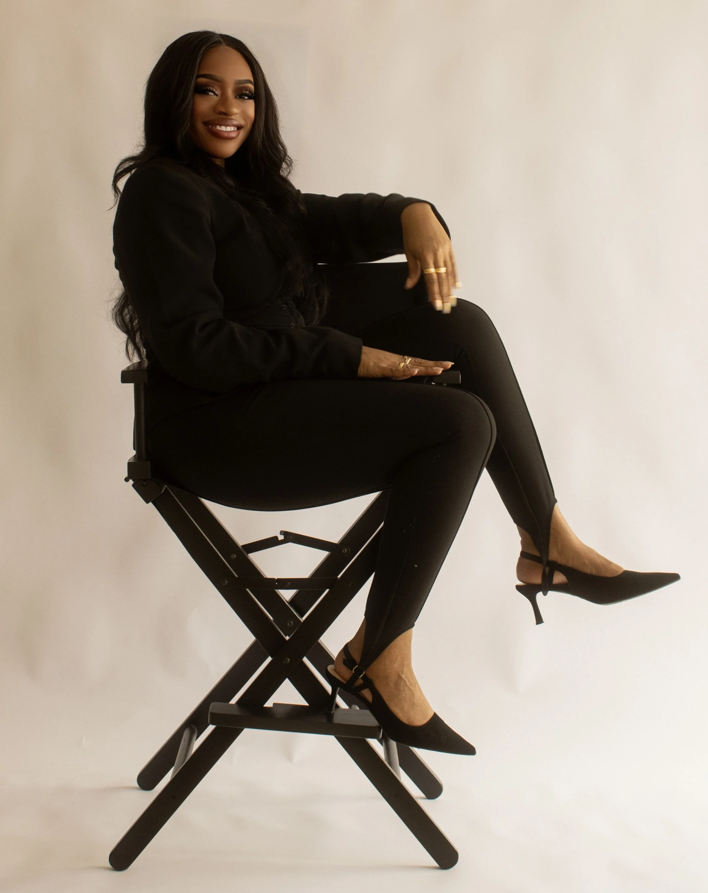 A woman with long dark hair and dark makeup sitting on a large black director's chair, wearing a black outfit and high heels, against a plain white background.