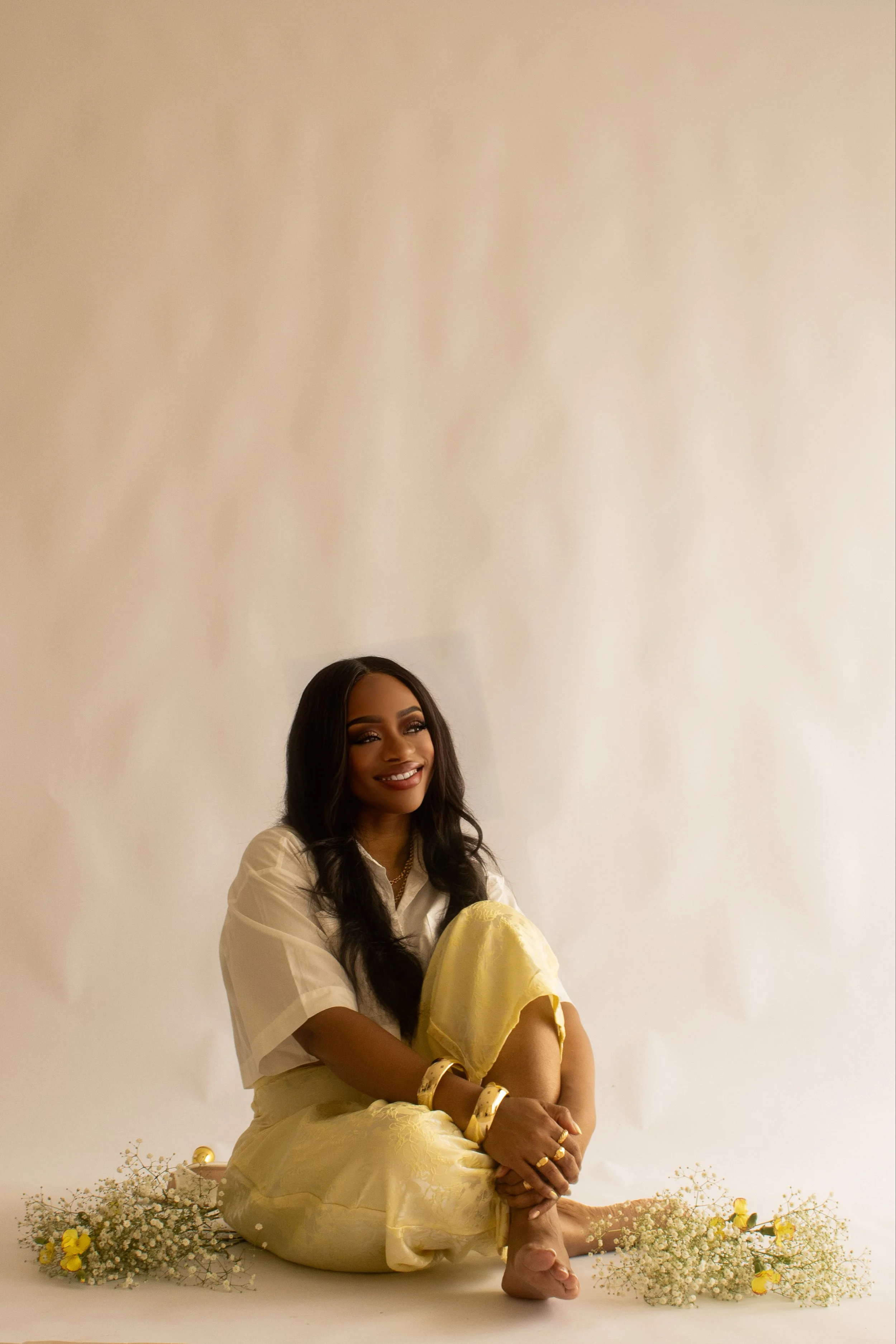 A woman with dark wavy hair and dark eyes sitting on the floor surrounded by white and yellow flowers, smiling and looking slightly to her left, against a plain beige background.