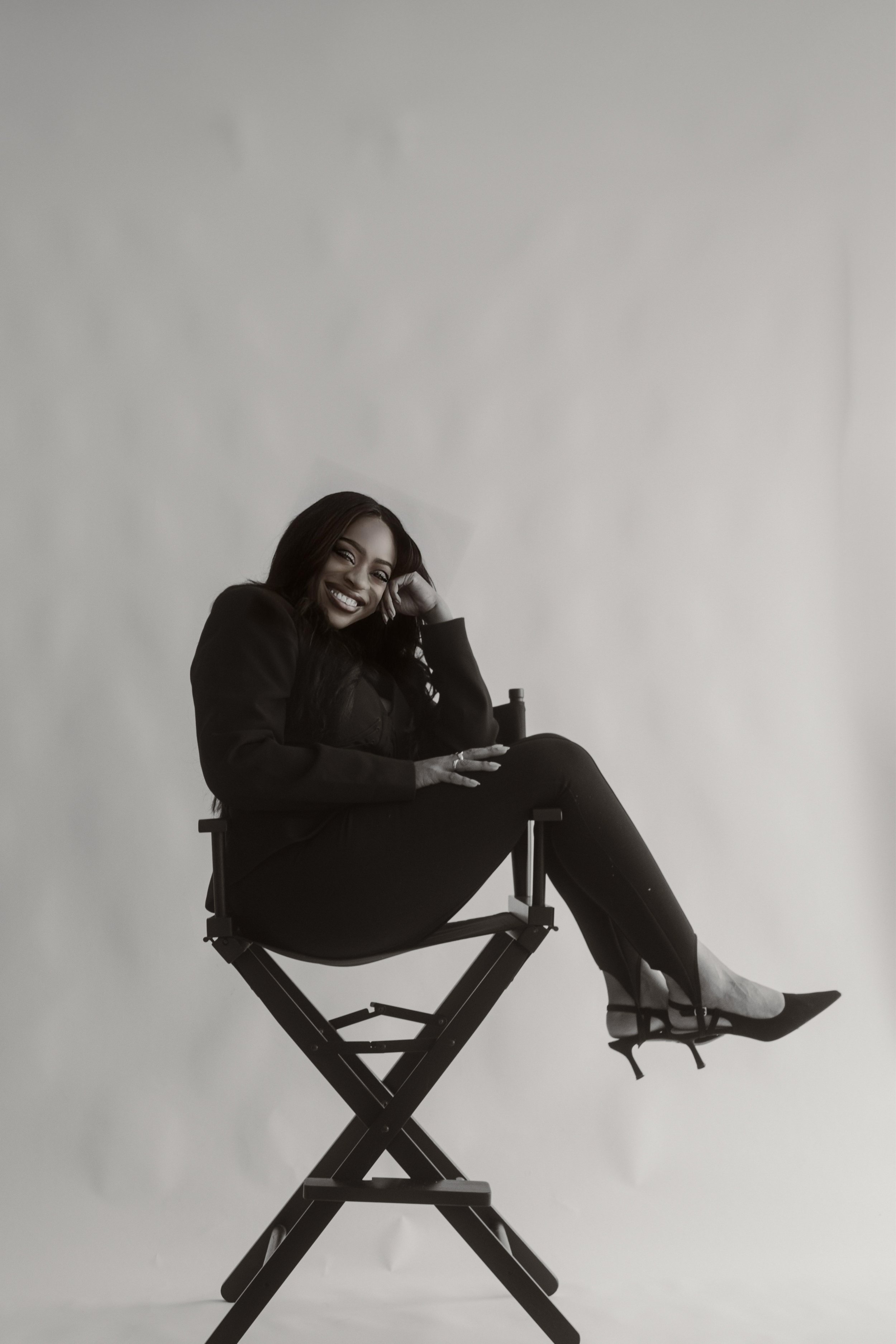 Black woman sitting on a director's chair, smiling, with her head resting on her hand, set against a plain light background.
