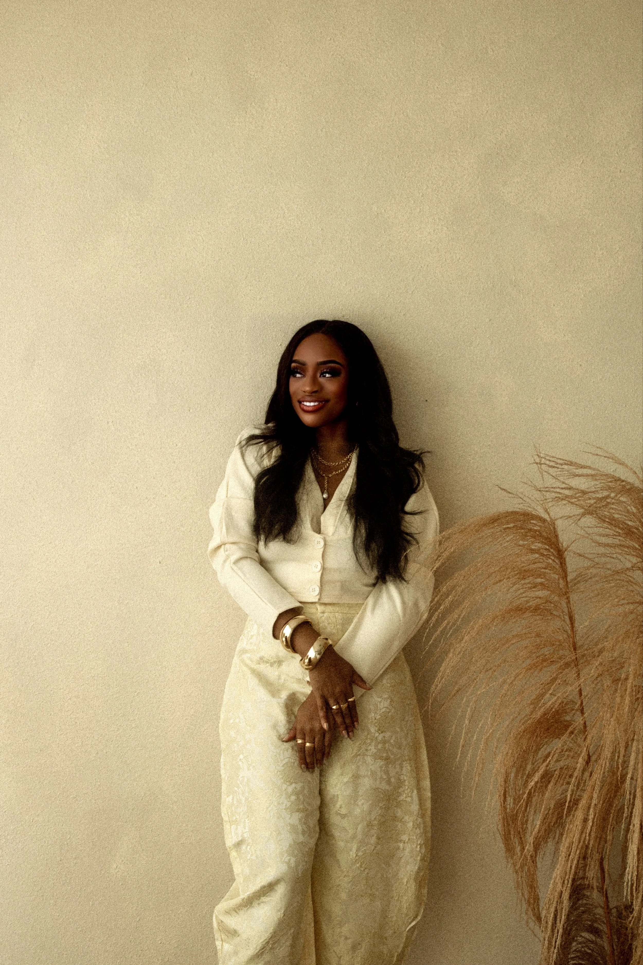 A woman with long, dark hair and jewelry, wearing a stylish cream-colored outfit, stands against a plain beige wall, with decorative dried plants visible on the right side of the image.