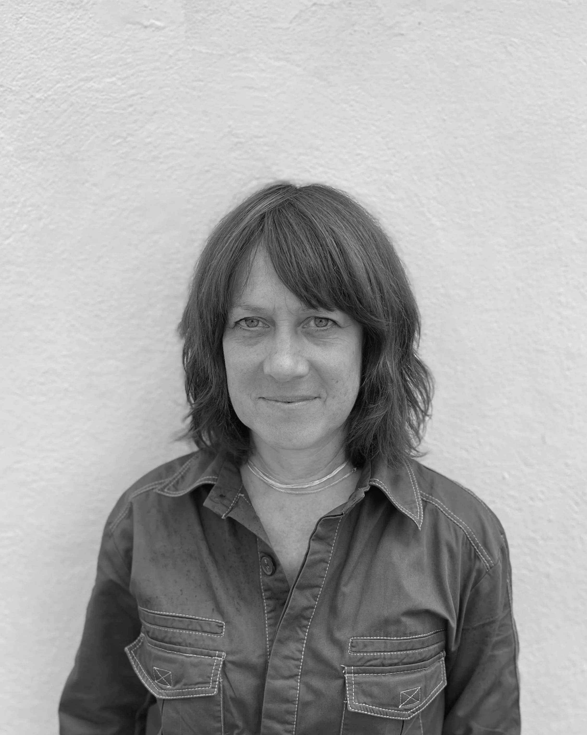 A woman with shoulder-length hair and light-colored eyes, wearing a button-up jacket, stands in front of a textured white wall.