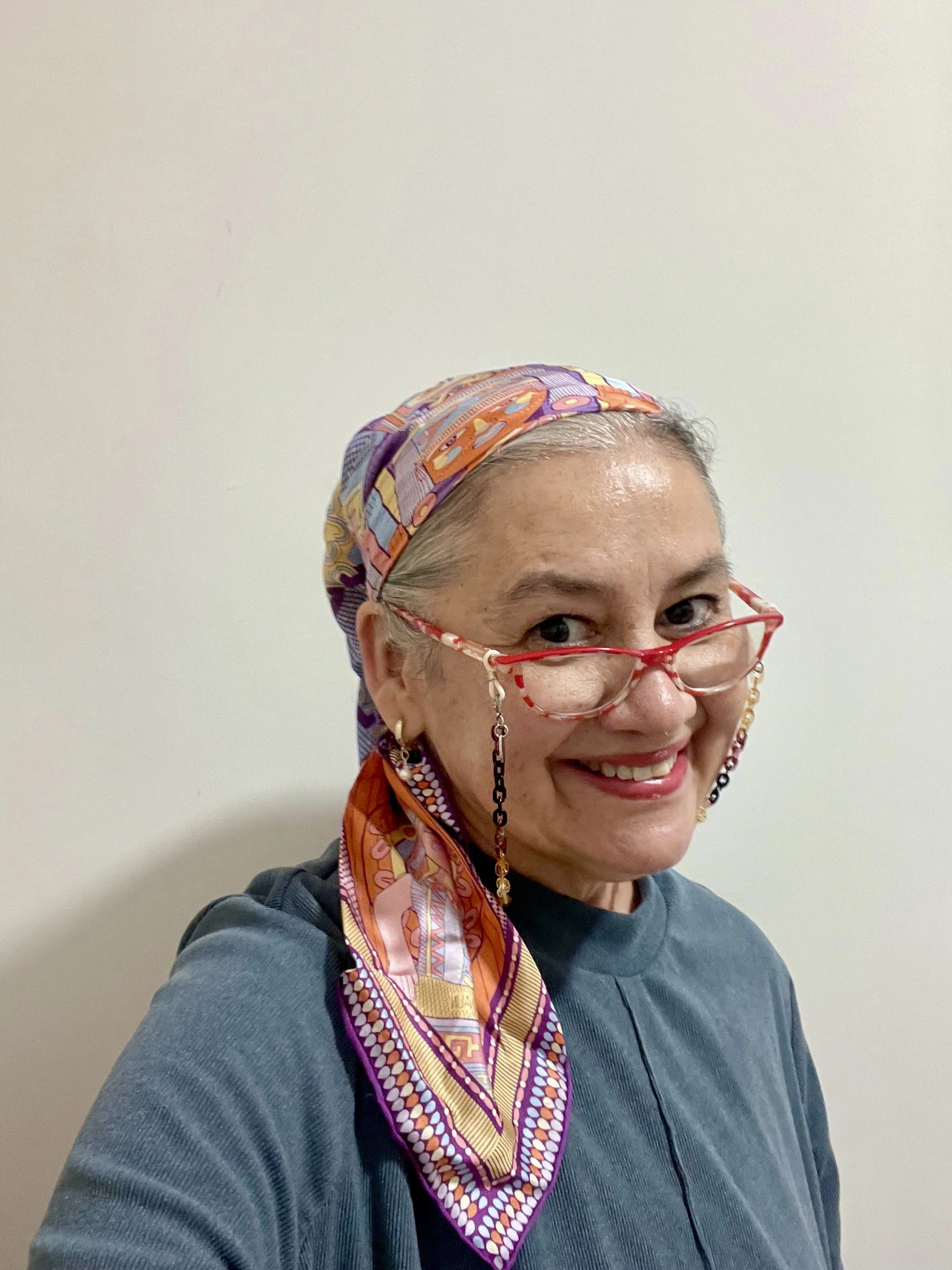 Mujer sonriente con gafas rojas y un pañuelo colorido en la cabeza, usando una camiseta azul y con un fondo blanco