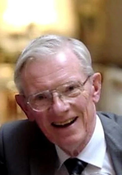 An elderly man with gray hair wearing glasses, a gray suit, and a white shirt, smiling and appearing to be in a social setting.