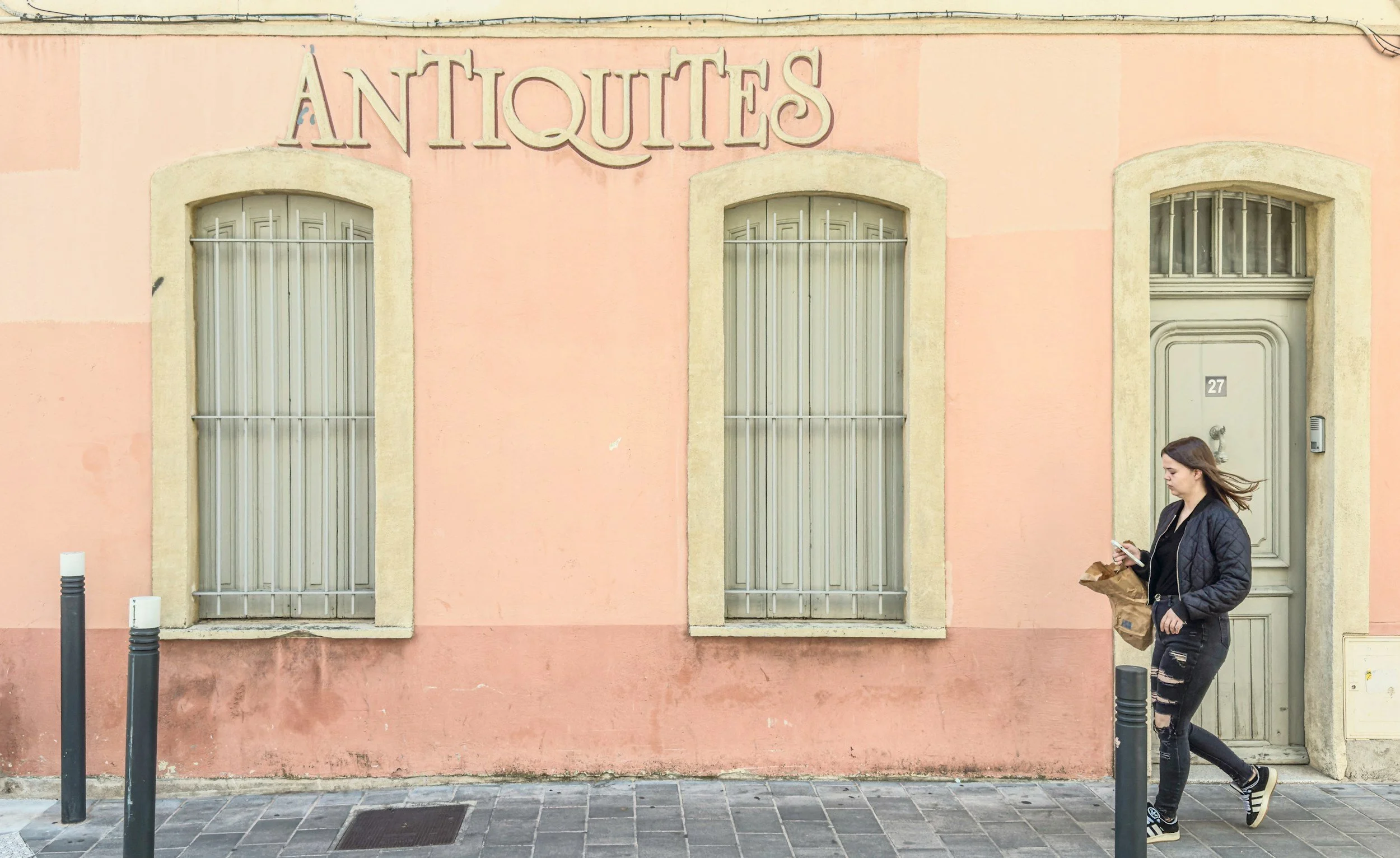 A woman walking past a pink building with barred windows and a door, holding a takeout bag and a smartphone.