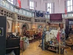 Interior of a vintage or thrift store with clothing racks, furniture, and displays, featuring a high ceiling and large windows.